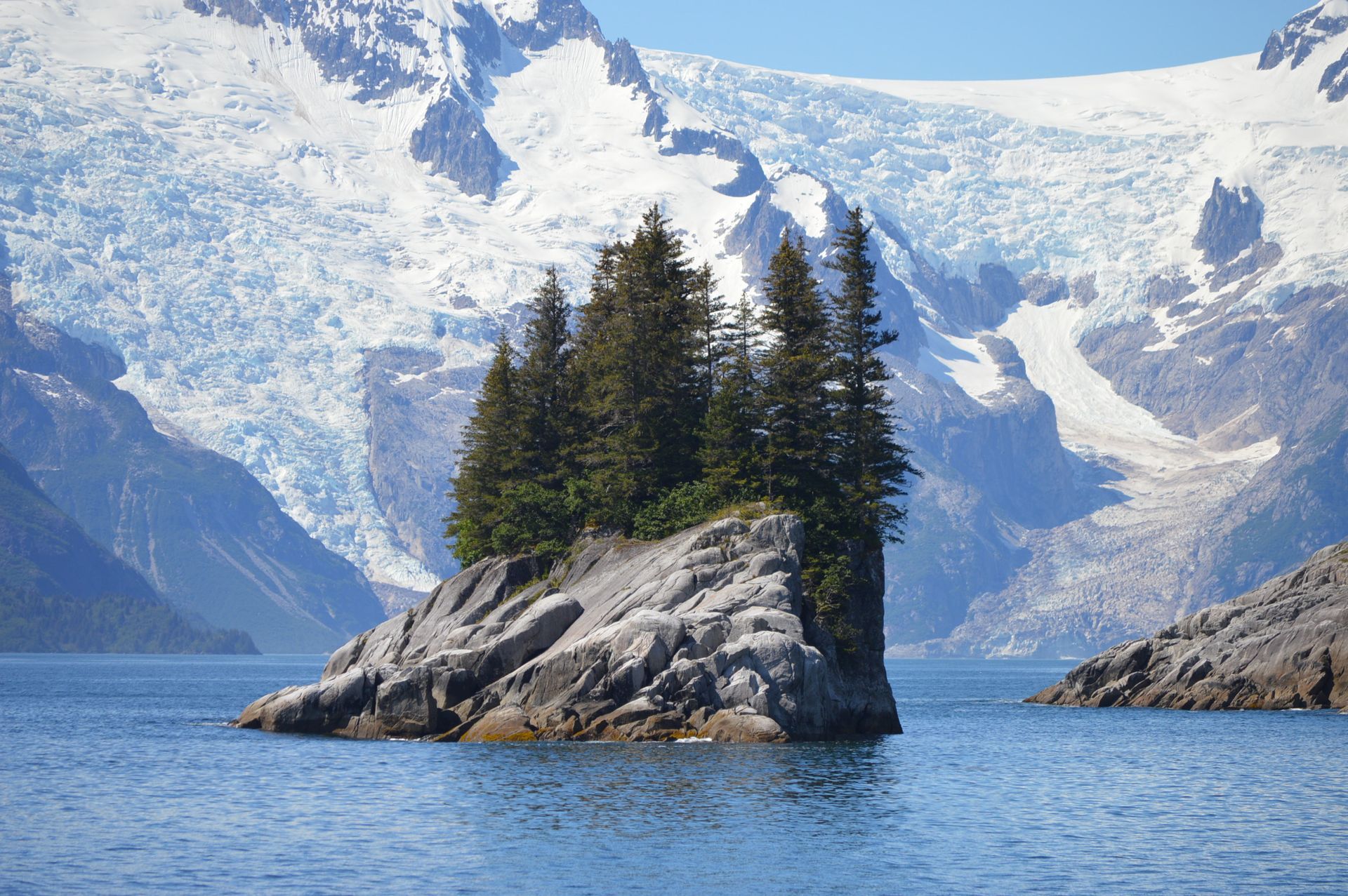 Rocky island with evergreen trees in front of a glacier and snow-covered mountains, in blue water.