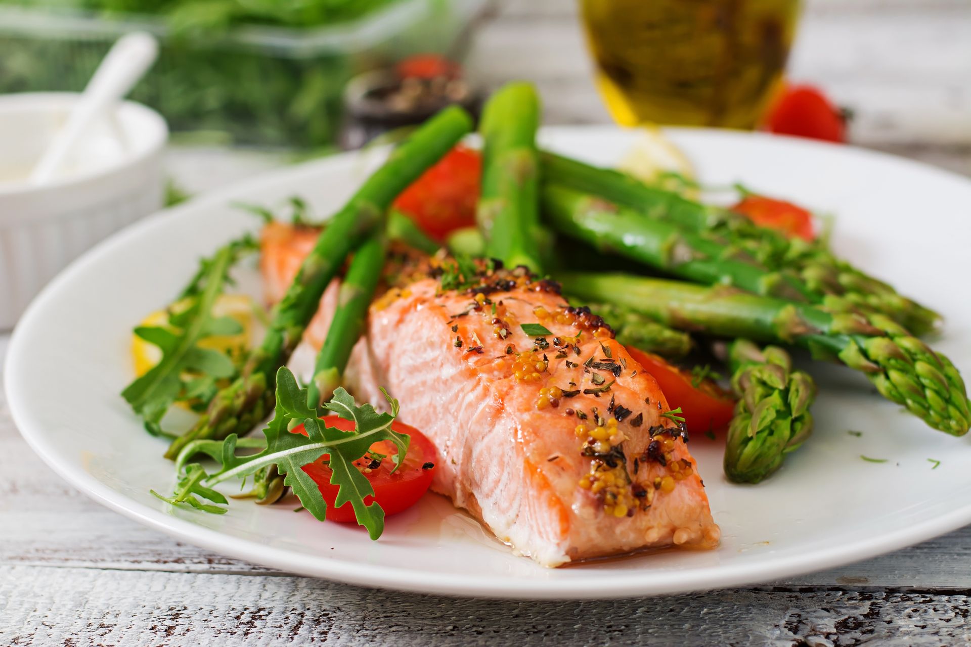 A plate featuring a seasoned salmon fillet, fresh asparagus, arugula, and cherry tomatoes on a rustic white wood table.