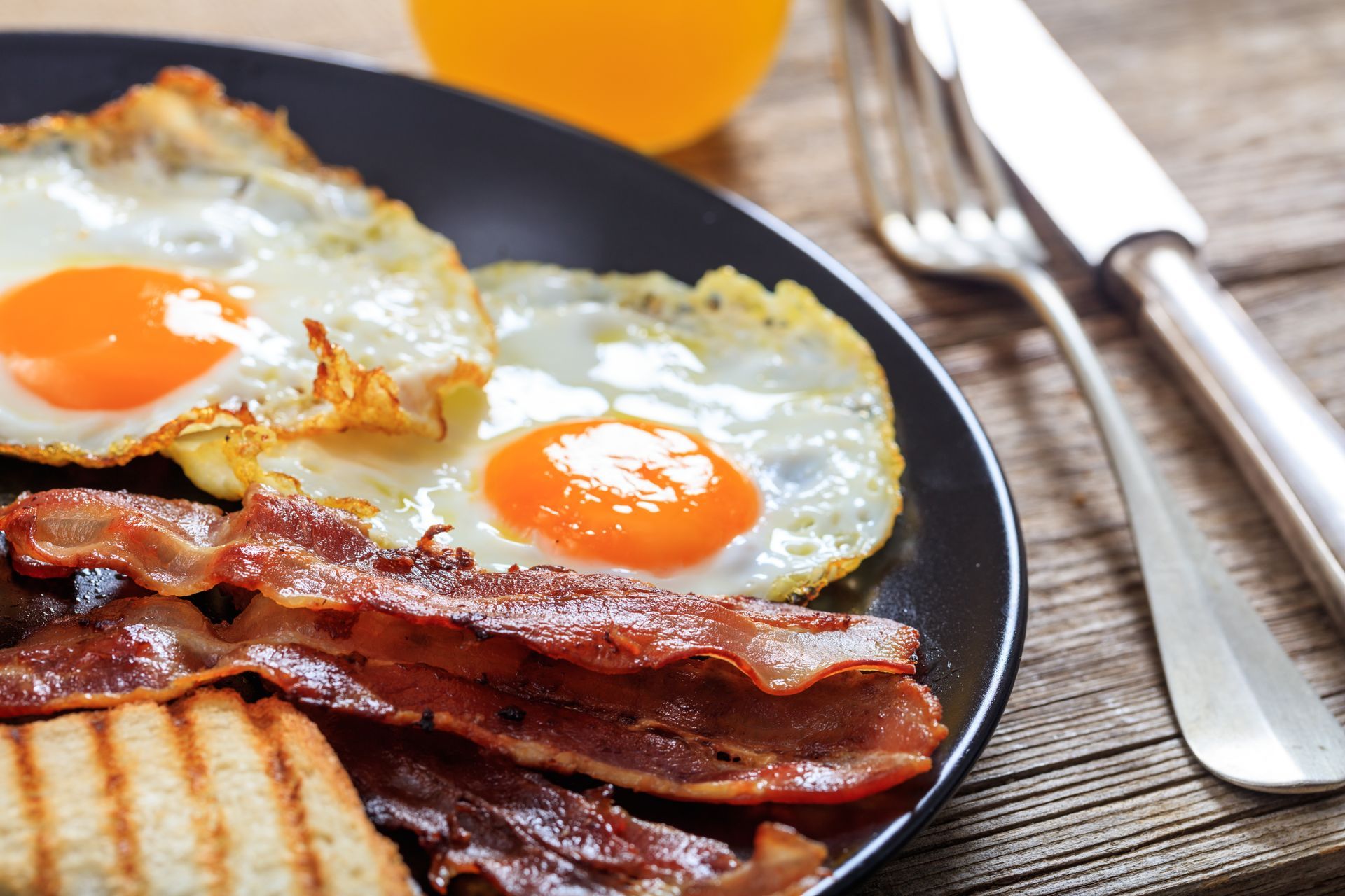 Fried eggs, crispy bacon, and grilled bread on a black plate, with a fork and knife on a wooden table.