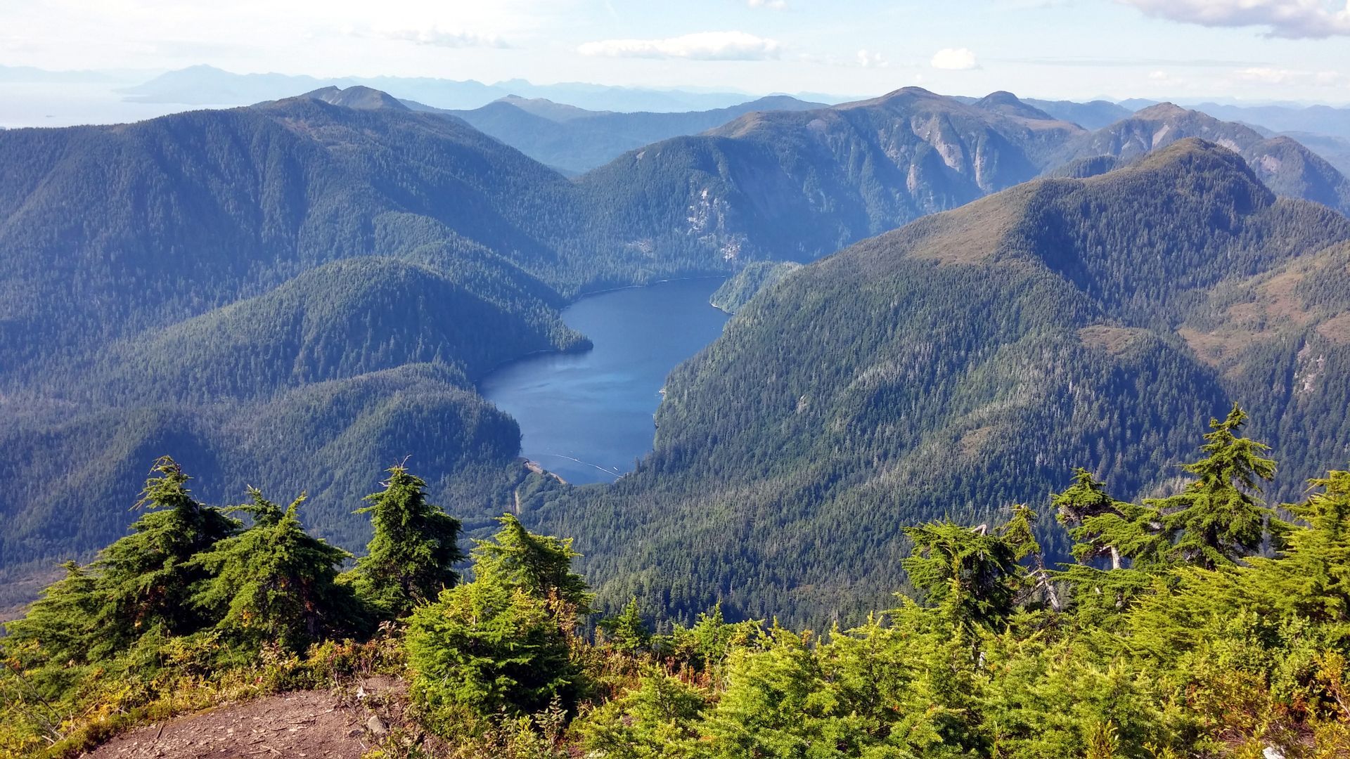 A vast, deep blue lake nestled between rugged, forested mountains under a partly cloudy sky.