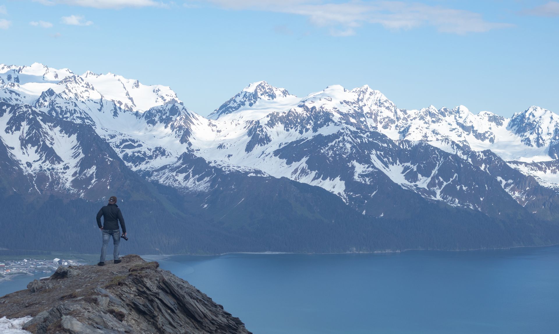 A person stands on a rocky mountain peak overlooking a deep blue bay and snow-capped mountain range under a clear sky.