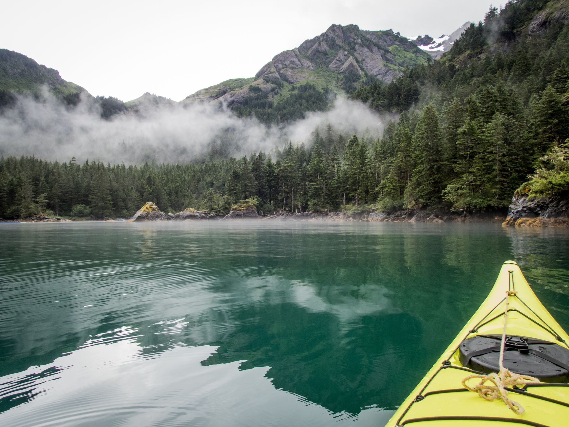 The bow of a bright yellow kayak pointing toward a calm, turquoise lake surrounded by misty pine forests and mountains.