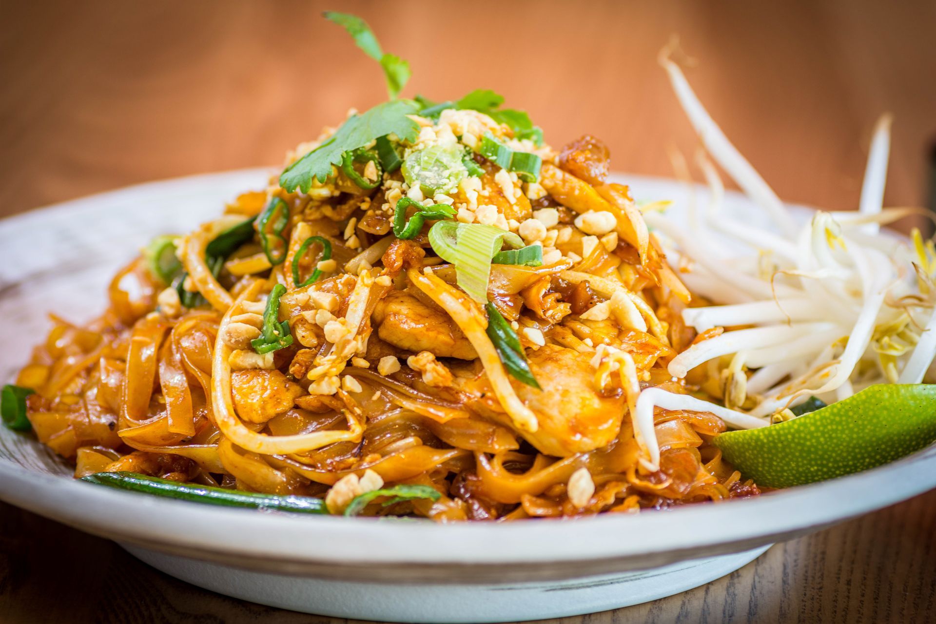 A plate of Pad Thai topped with crushed peanuts and green onions, served with a lime wedge and fresh bean sprouts.