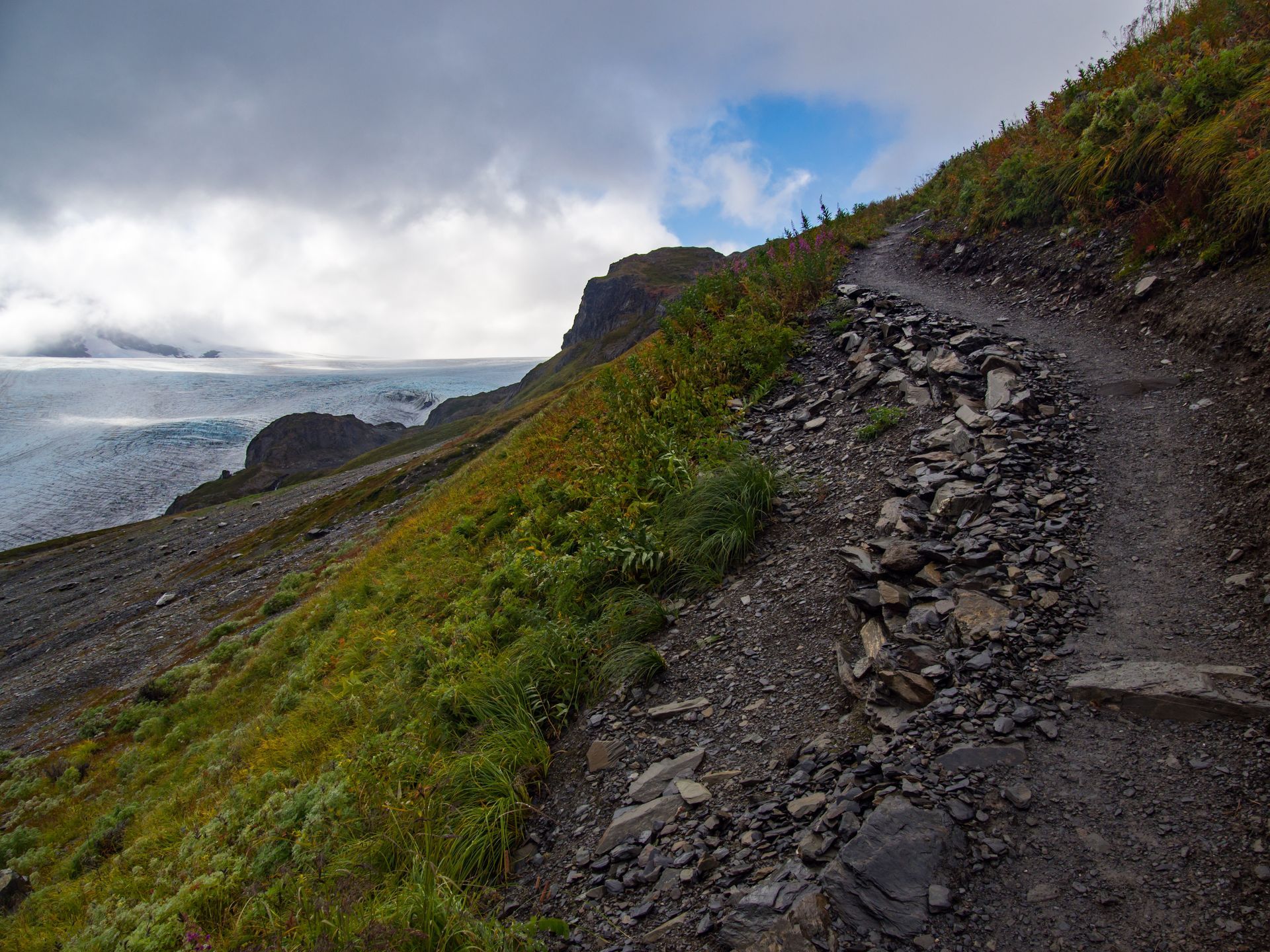 A narrow, rocky hiking trail winds along a steep, grassy mountainside overlooking a vast, cloudy glacier.