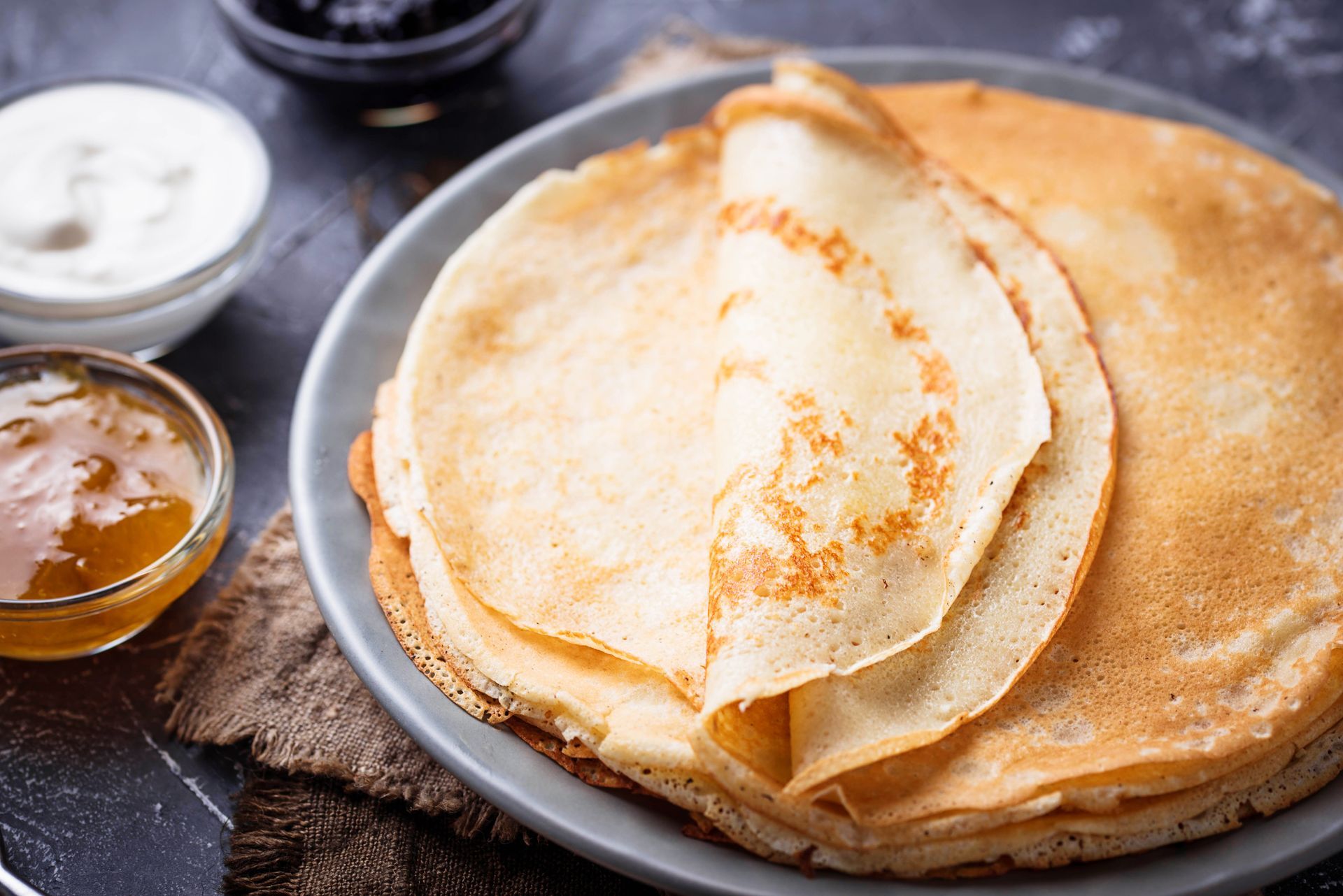A stack of thin, golden-brown crepes on a plate, served with small bowls of jam and sour cream on a dark surface.