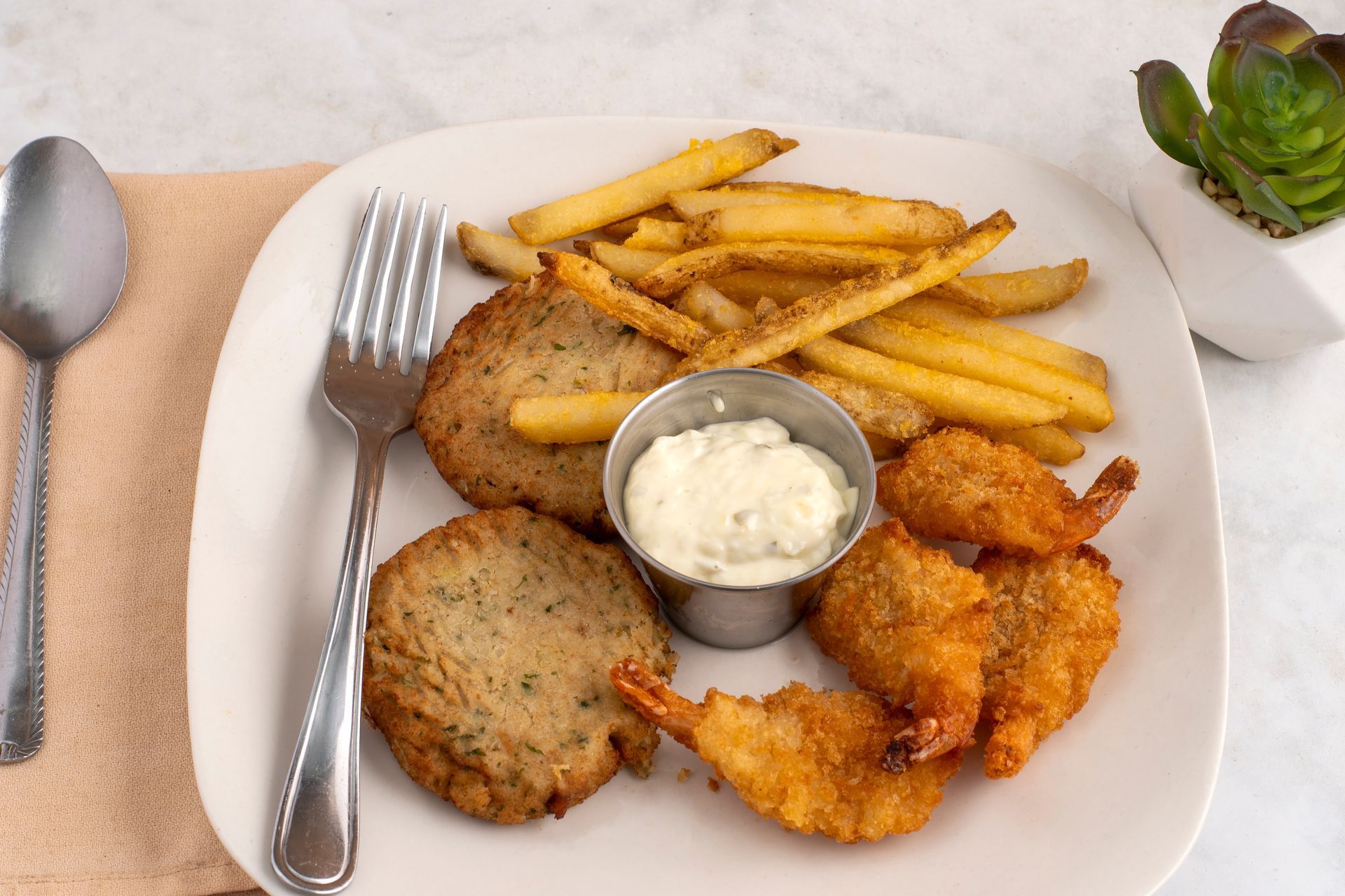 A square plate featuring two crab cakes, fried shrimp, French fries, and a small cup of dipping sauce, with a fork nearby.