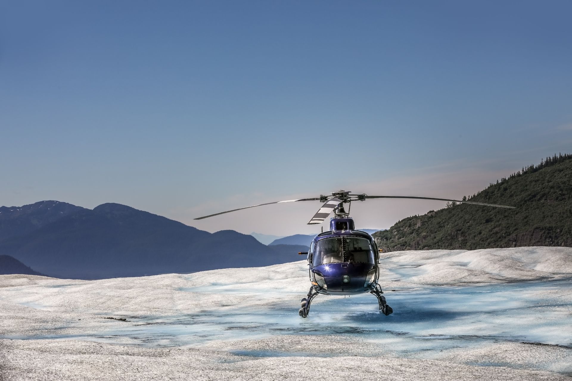 A blue helicopter parked on a bright, icy glacier with distant mountains under a clear blue sky.