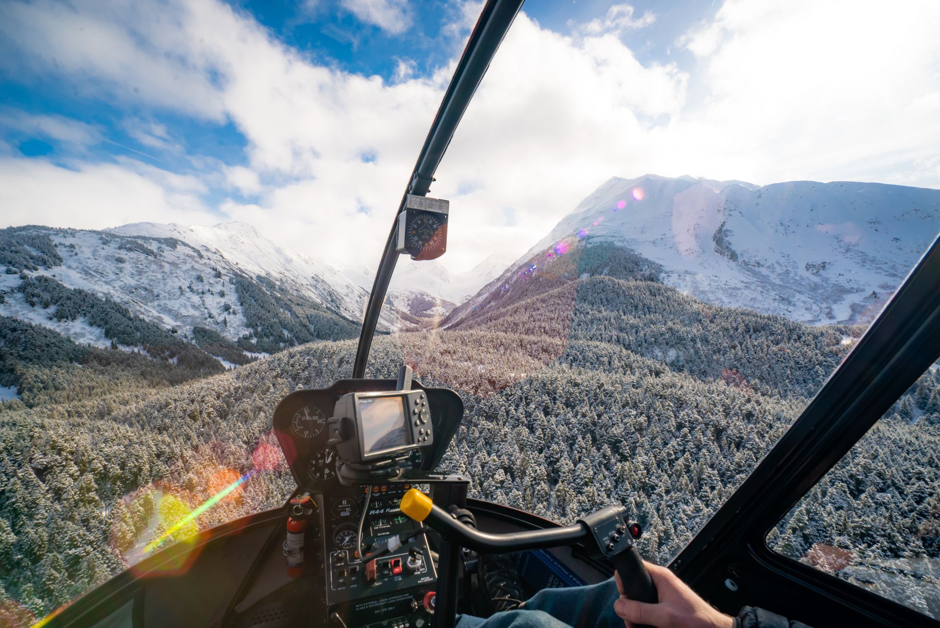 A first-person view from a helicopter cockpit flying over a snow-covered mountain forest under a bright, sunny sky.
