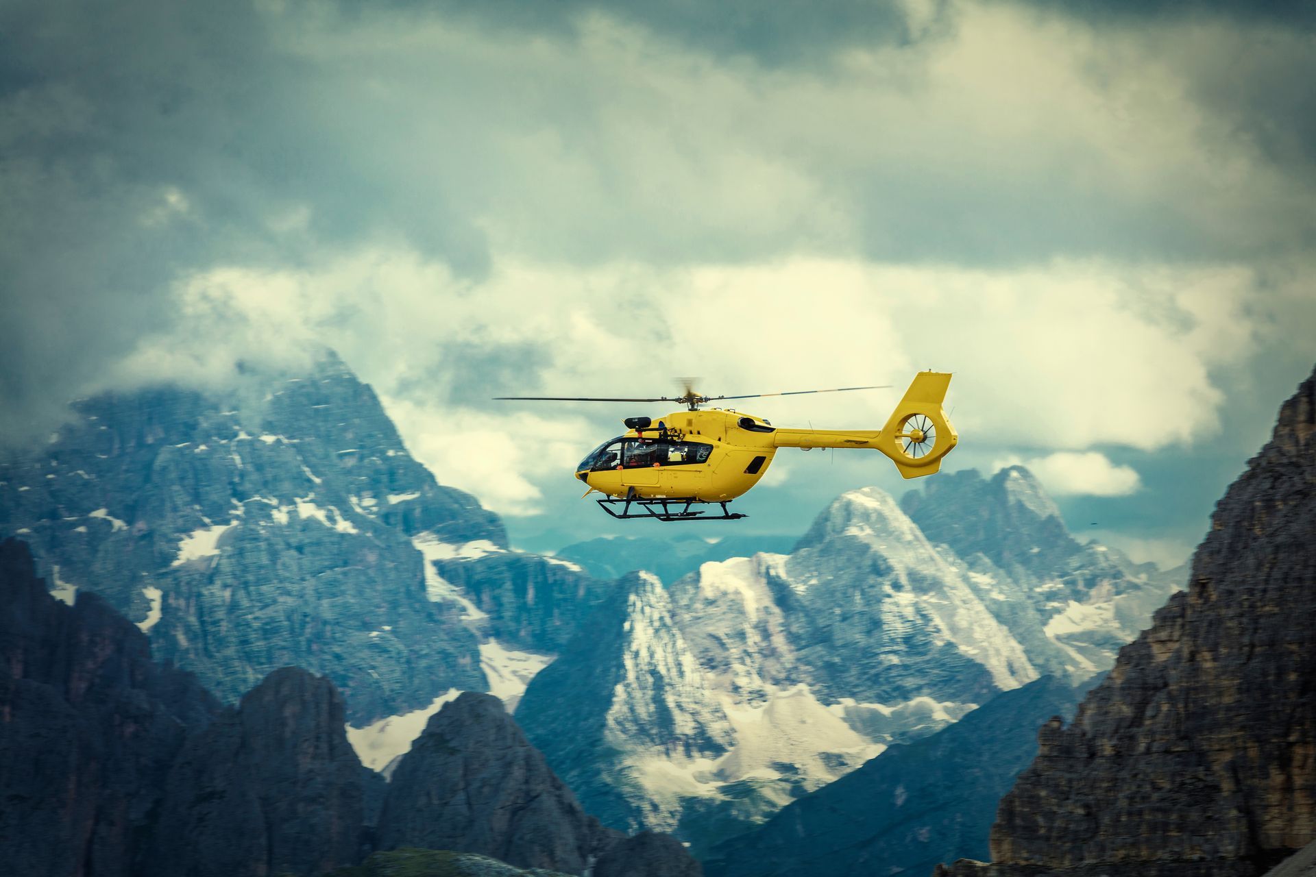 A yellow rescue helicopter flies through dramatic, dark, cloudy skies above rugged, snow-dusted mountain peaks.