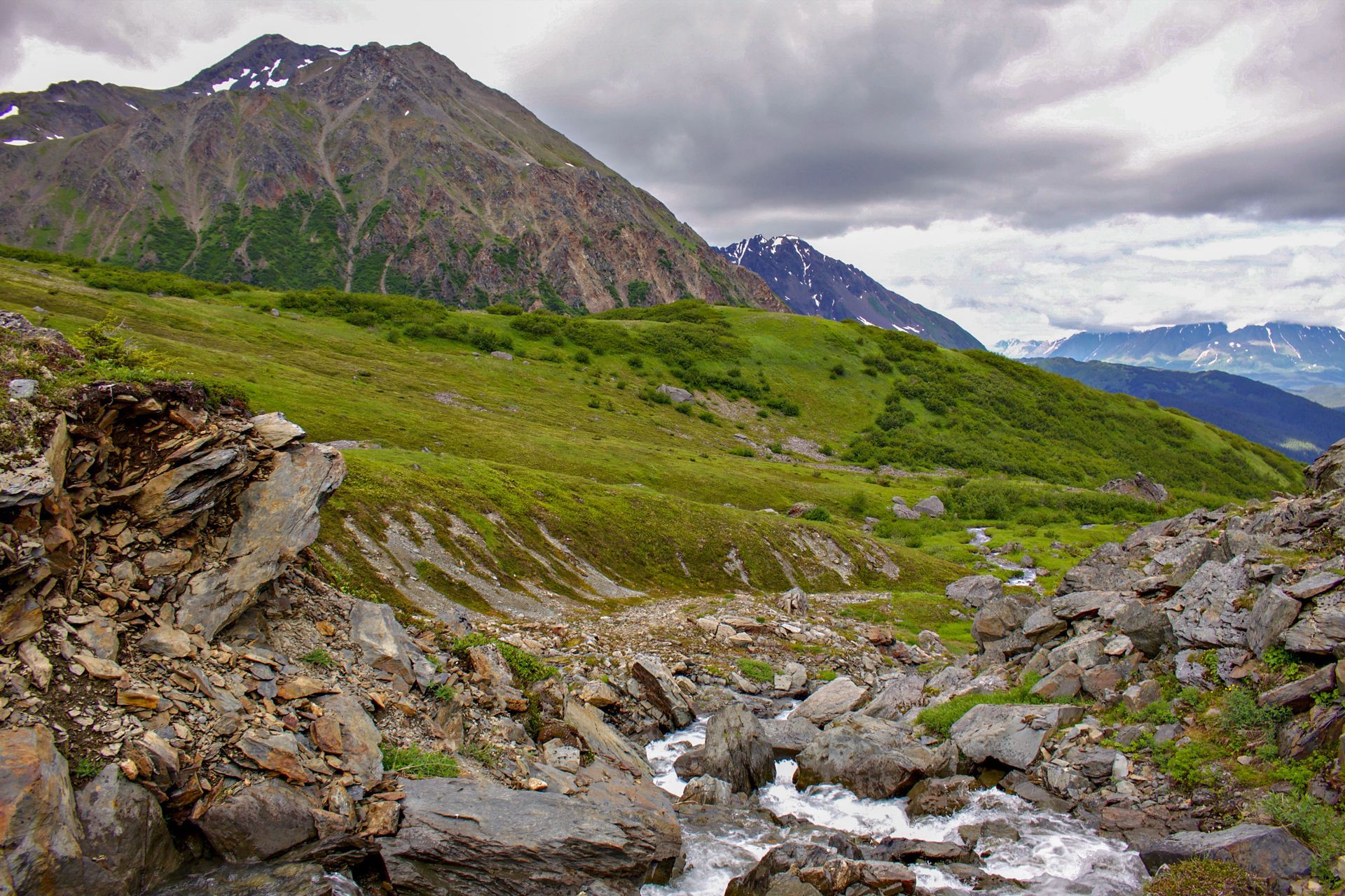 A mountain stream flows through a rocky valley under a cloudy sky, with green slopes rising toward jagged peaks.