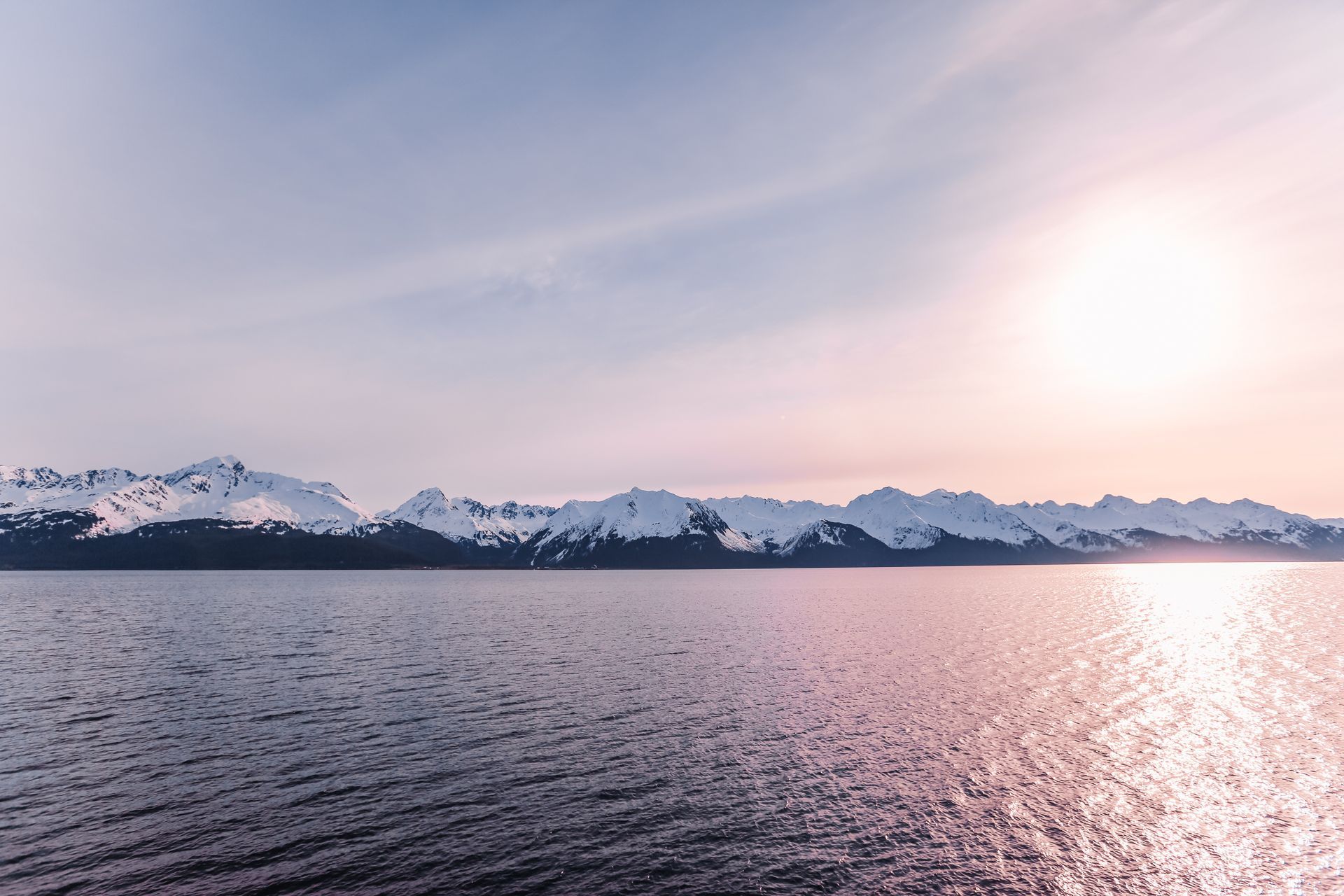 Sunlight reflects on calm water before a distant, snow-capped mountain range under a soft, pastel-toned sky.