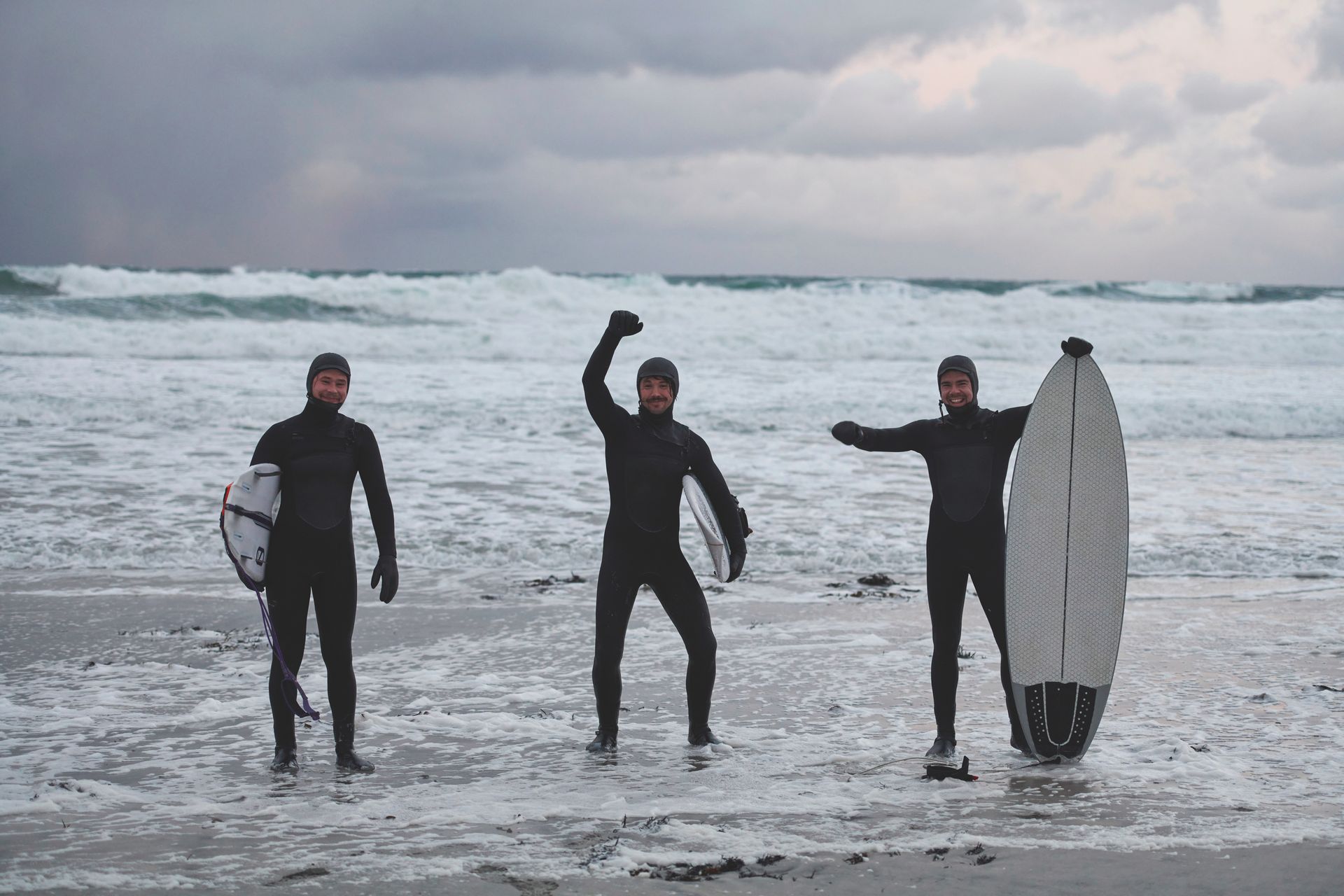 Three surfers in wetsuits pose on a sandy beach by a wavy ocean under a cloudy sky, with one holding a surfboard aloft.