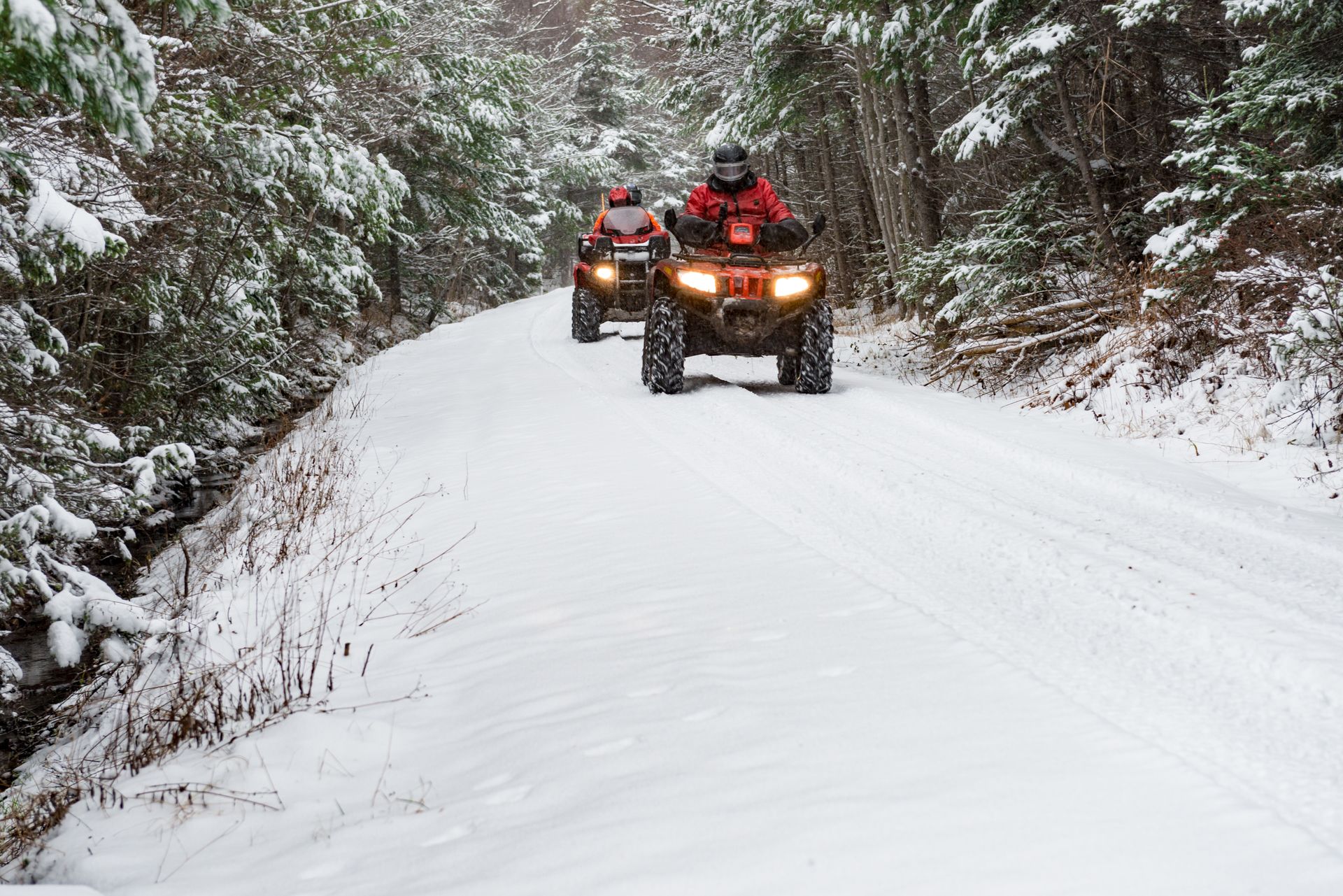 Two ATVs with headlights on drive along a snowy forest trail