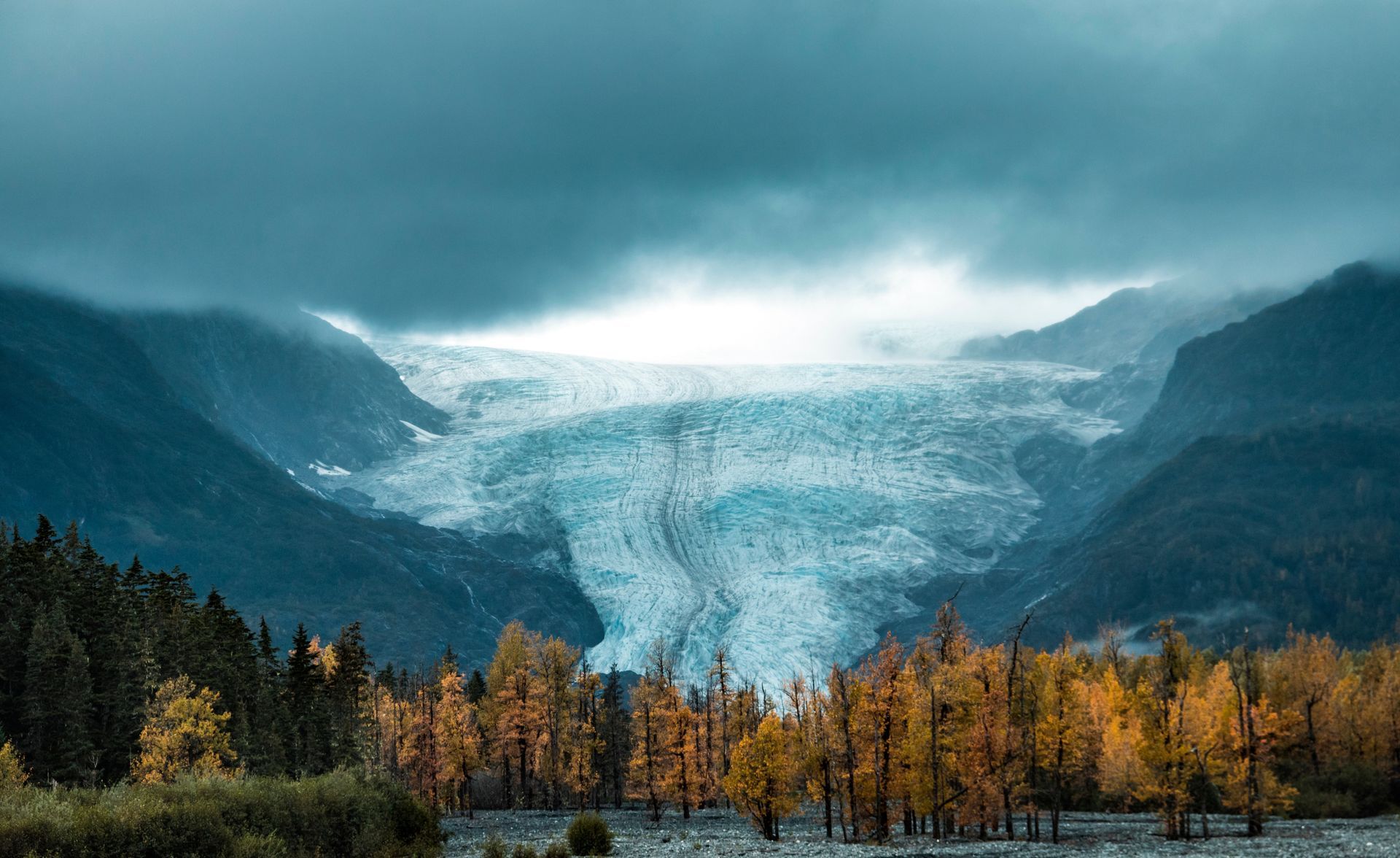 A large blue glacier flows down between dark mountain slopes above a forest with golden autumn foliage.