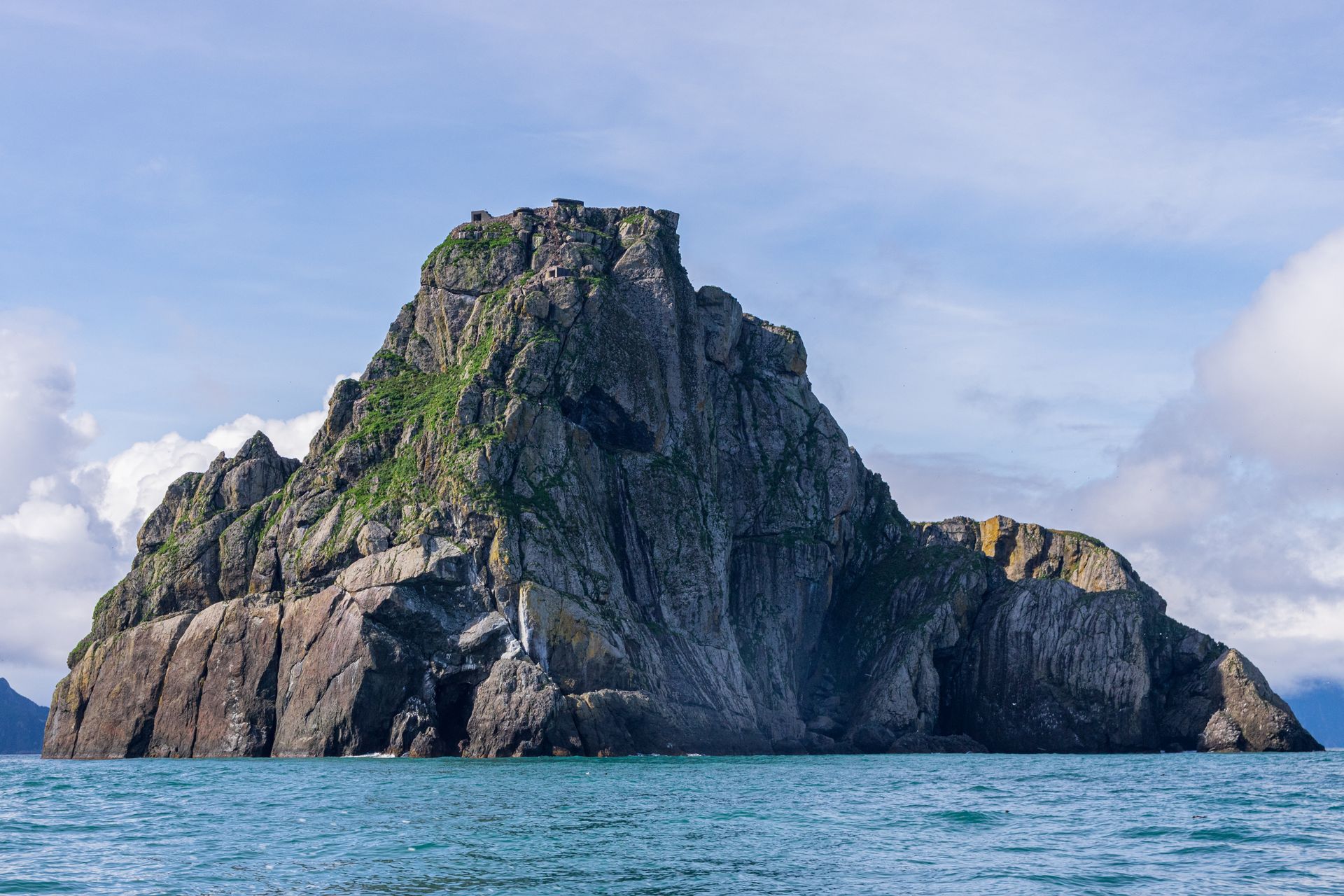 A craggy, steep-sided rock island rising from deep blue waters under a pale blue sky with light clouds.