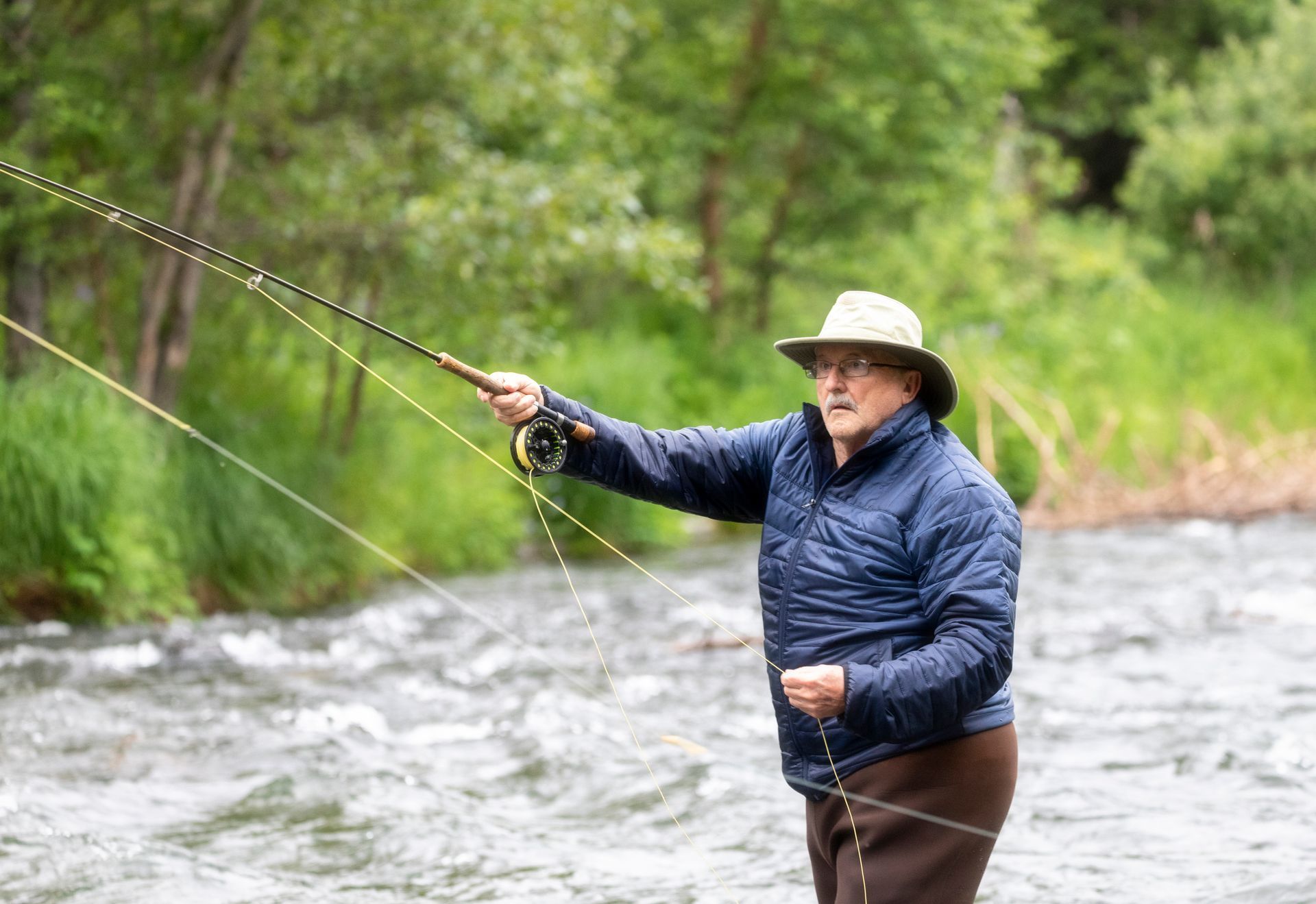 An angler wearing a sun hat and blue jacket fly-fishes in a rushing river surrounded by trees.