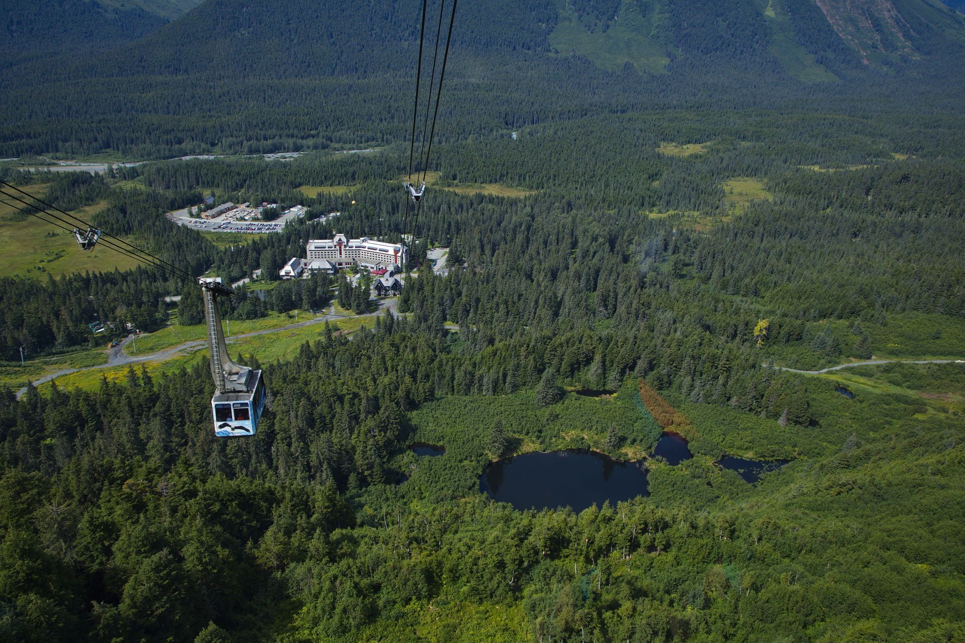 An aerial tram car descends over a lush, forested landscape with a small lake and a building complex in the distance.