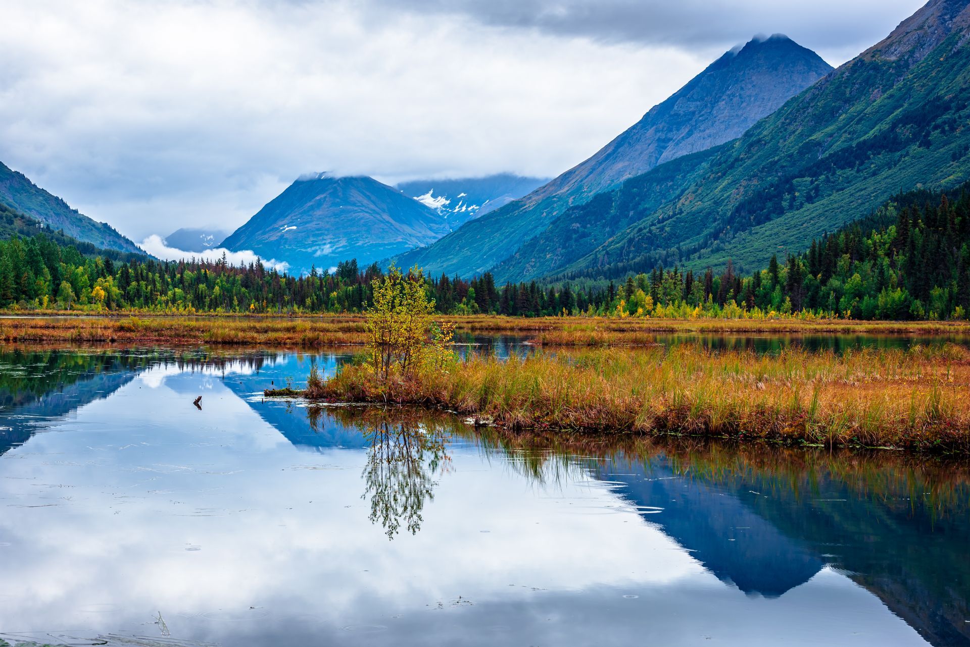 A calm lake reflects towering, rugged mountains and a line of evergreen trees under a cloudy sky.