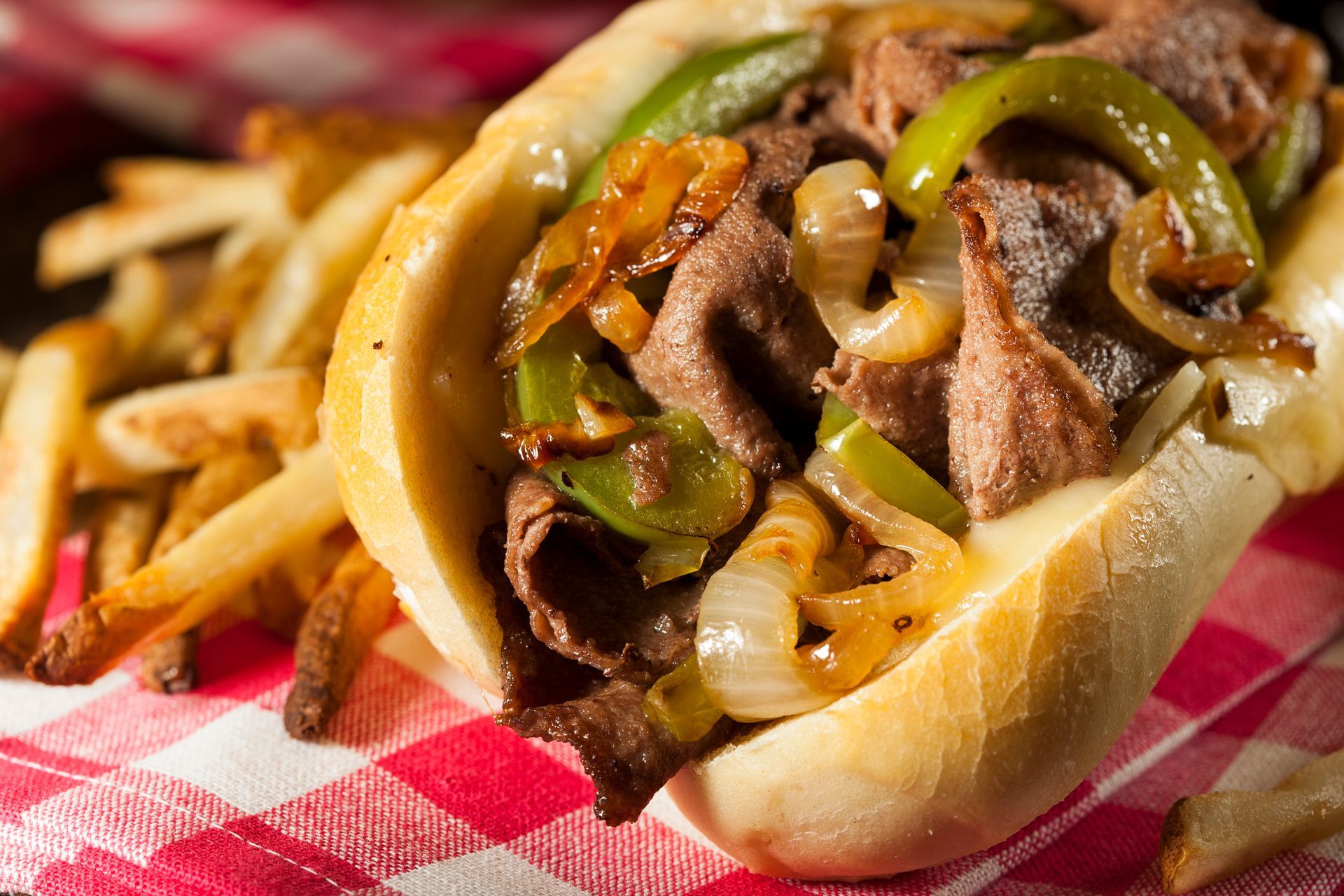 A Philly cheesesteak sandwich topped with peppers and onions, served with french fries on a red-and-white checkered cloth.