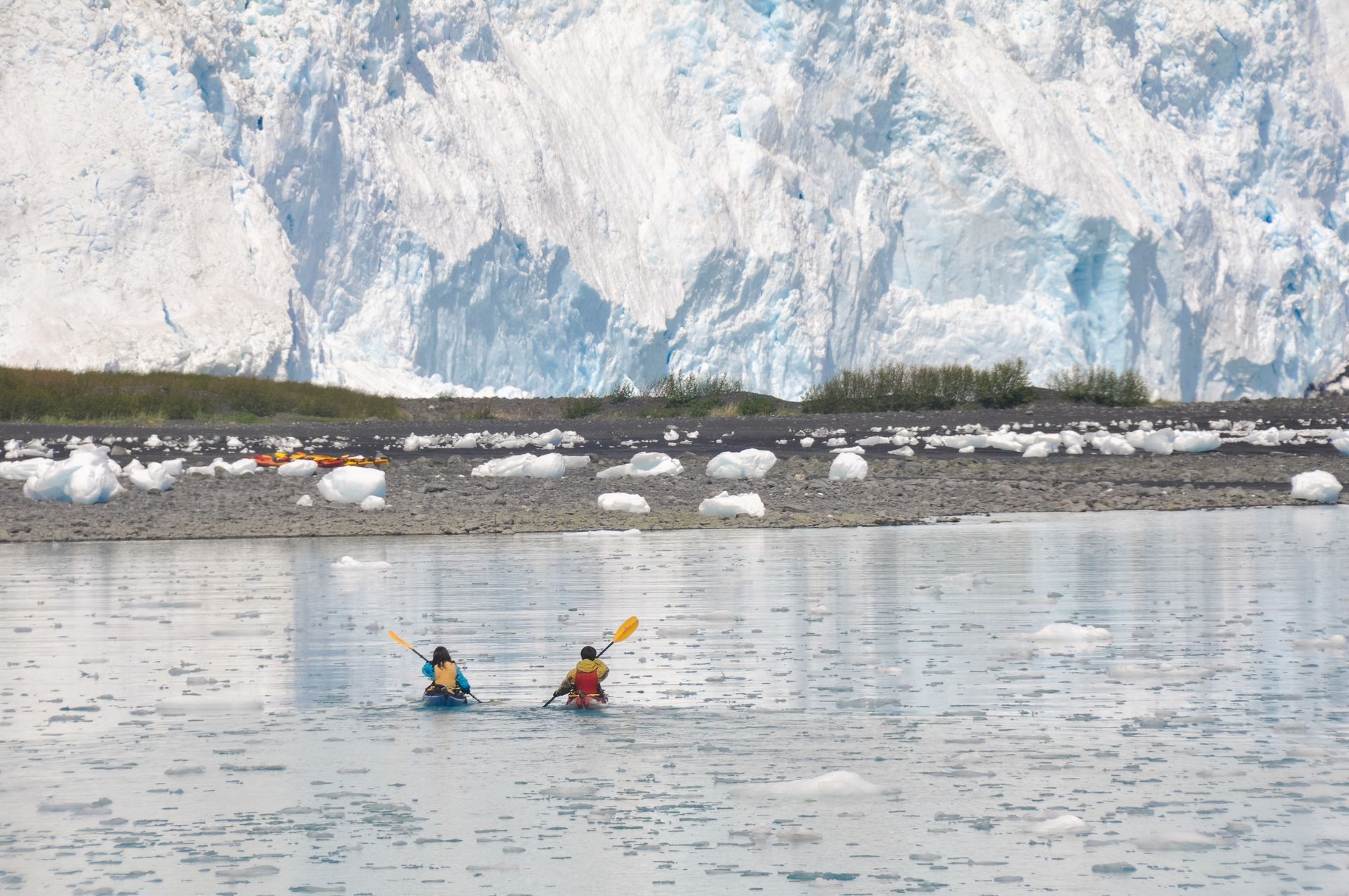 Two people in kayaks paddle through icy, calm water in front of a rocky shoreline and a massive, towering glacier.