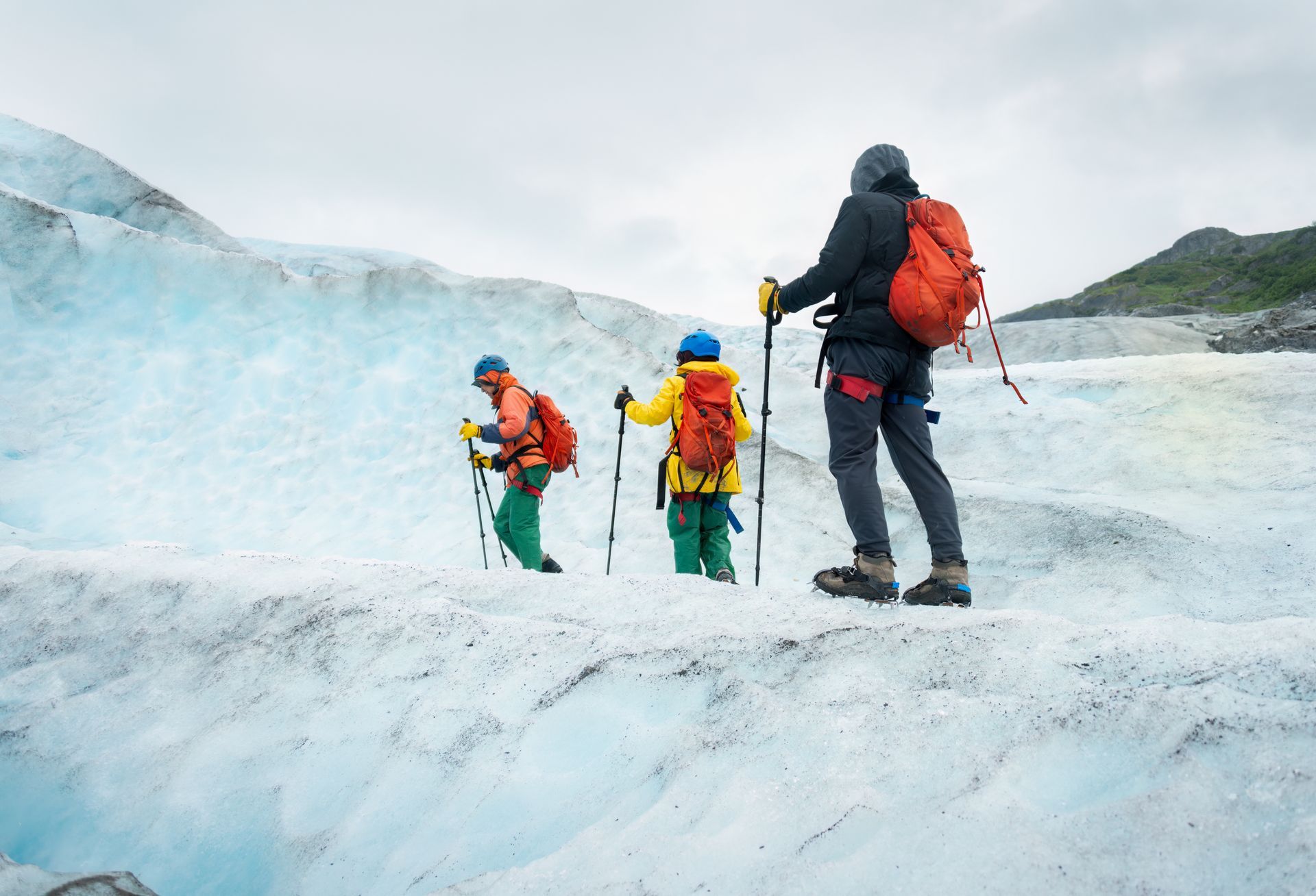 Three people in hiking gear and bright backpacks walk across a glacier, using trekking poles for balance.