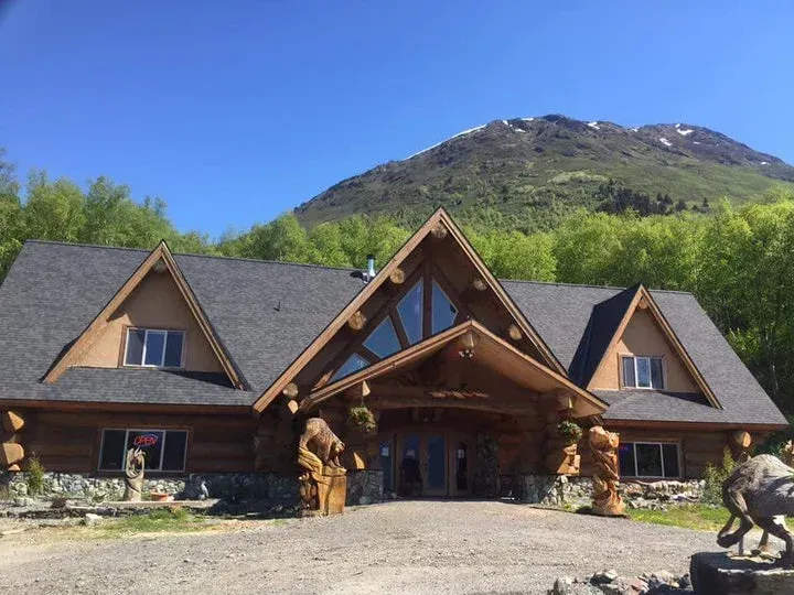 A log cabin lodge with a large peaked roof, stone base, and wooden carvings sits before a lush green mountain.