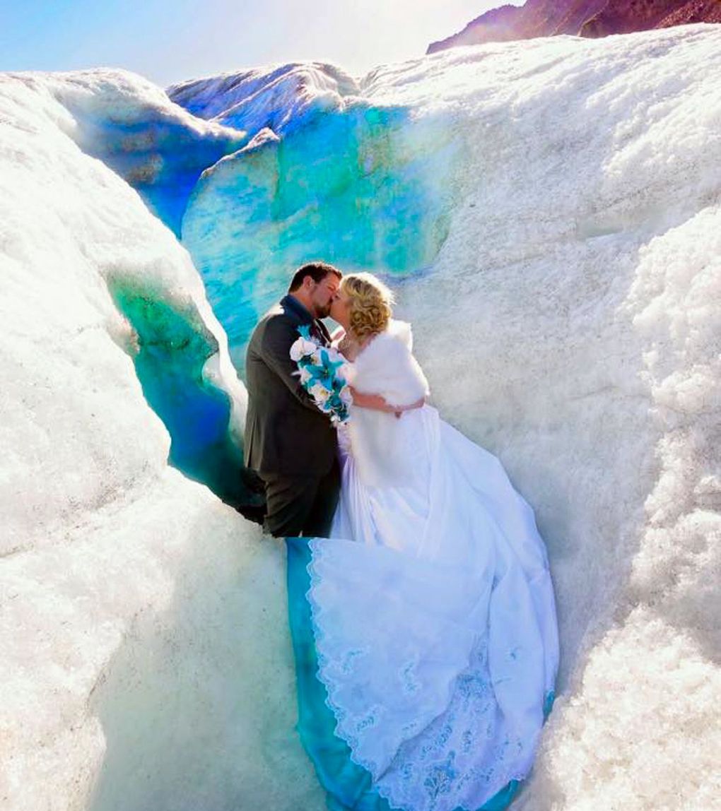 Couple kissing in a glacier crevice; bride in white dress, groom in suit, turquoise ice.