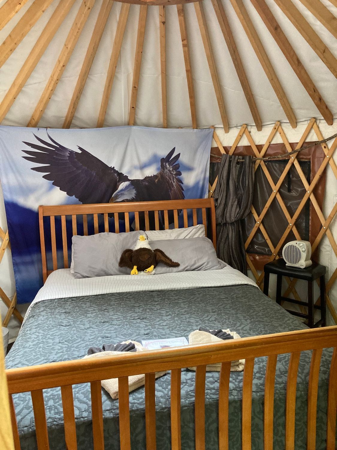 Inside a yurt, a bed with eagle decor: tapestry, pillow, stuffed bird. Brown wood frame, window, and a small table.