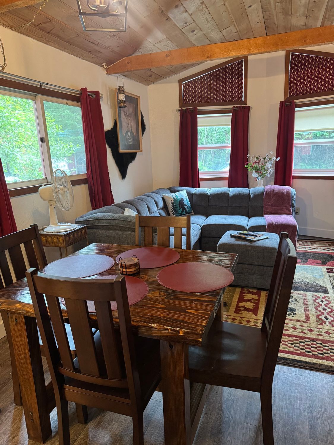 Dining area with table and chairs, adjacent to a sectional sofa. Windows with red curtains.