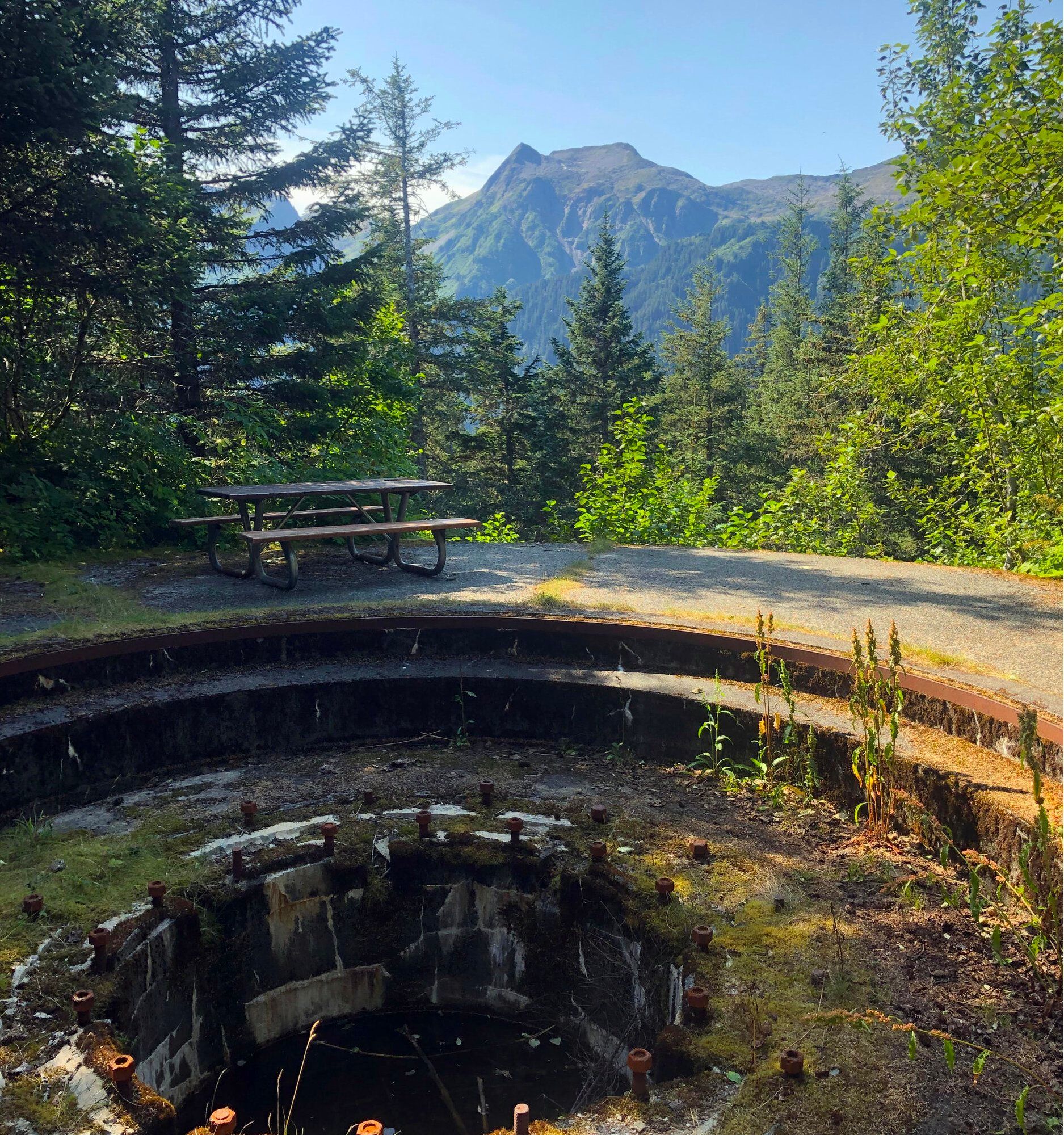 A picnic table sits on a gravel overlook next to a stone artillery gun emplacement, backed by pine trees and mountains.