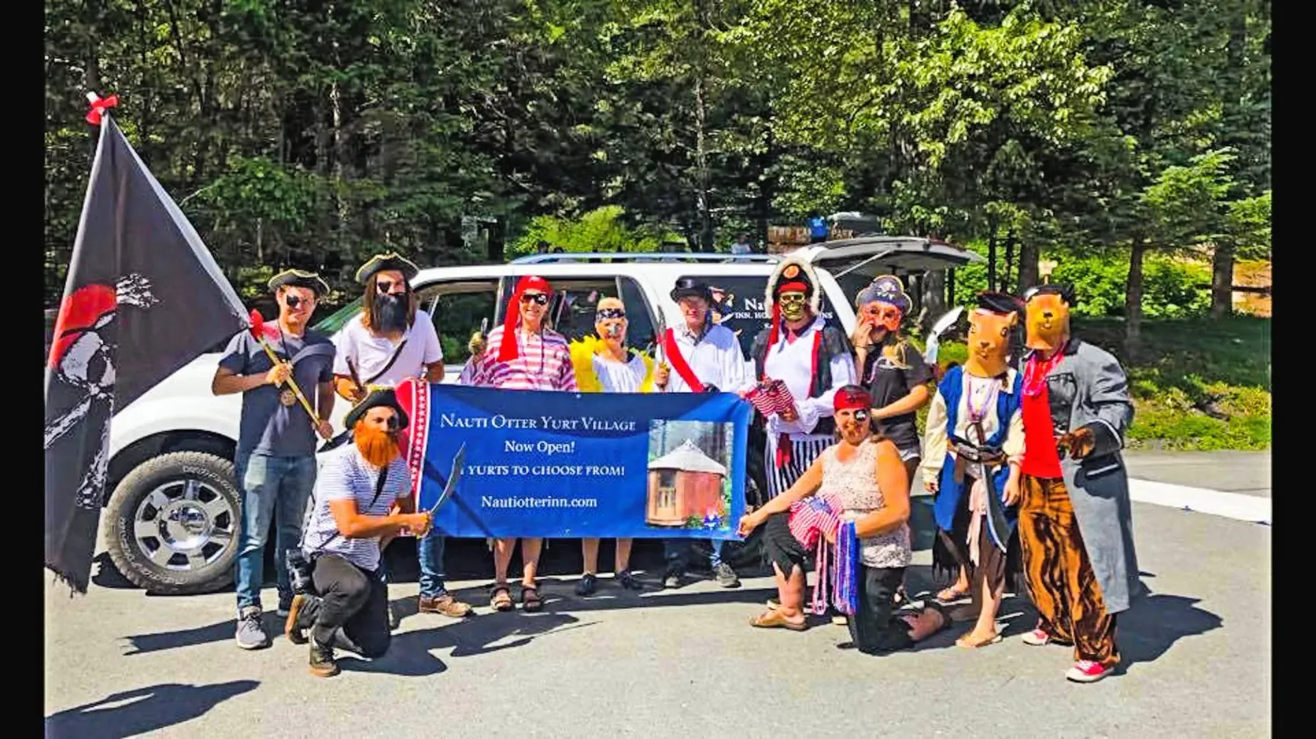 Group in pirate costumes holding a banner in front of a van.