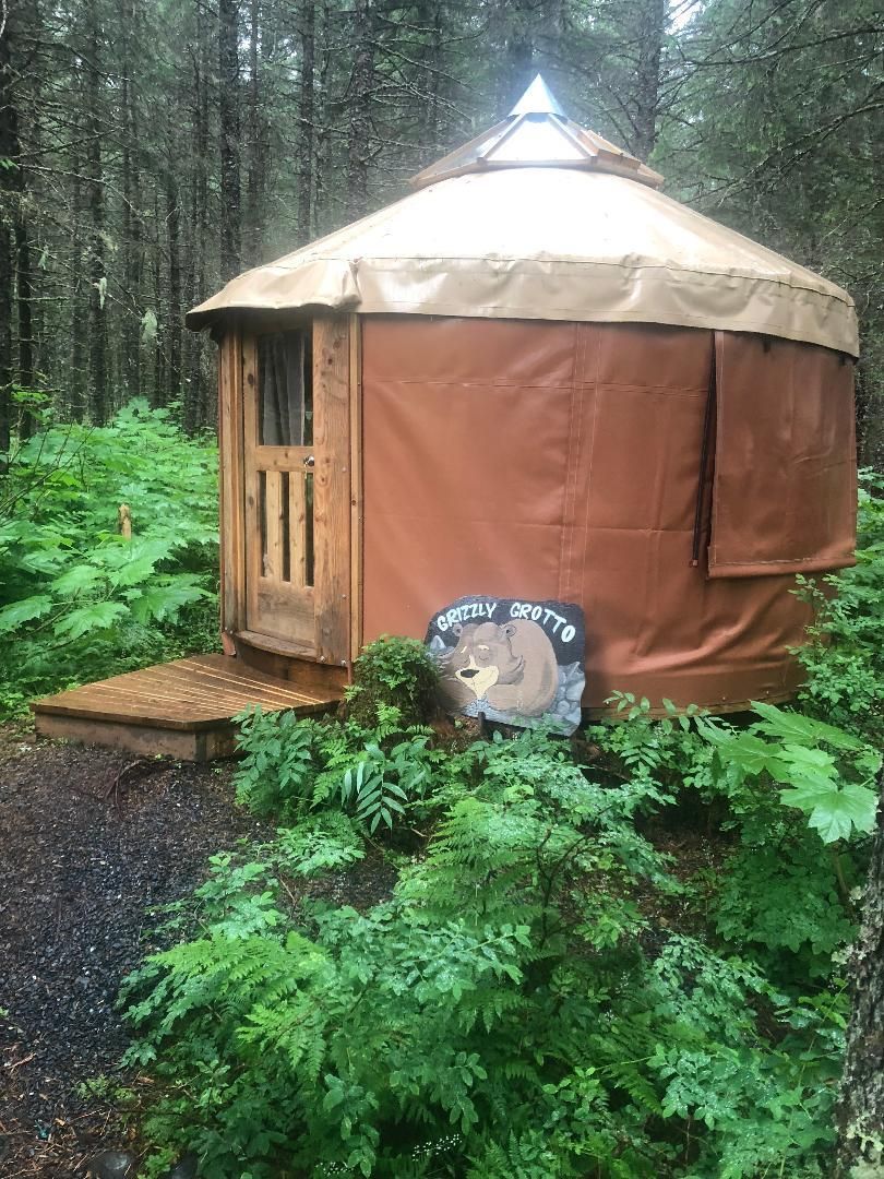 Yurt in a forest clearing; brown canvas exterior, wooden door, surrounded by lush green plants.
