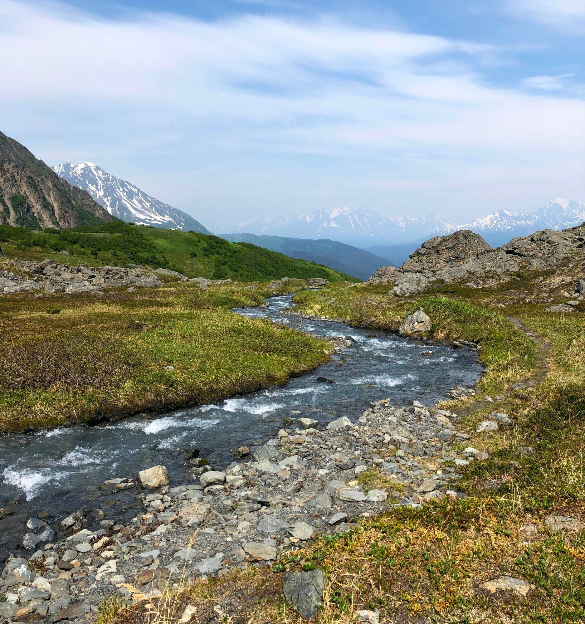 A clear, rocky stream flows through a lush, green mountain valley towards distant, snow-capped peaks under a blue sky.