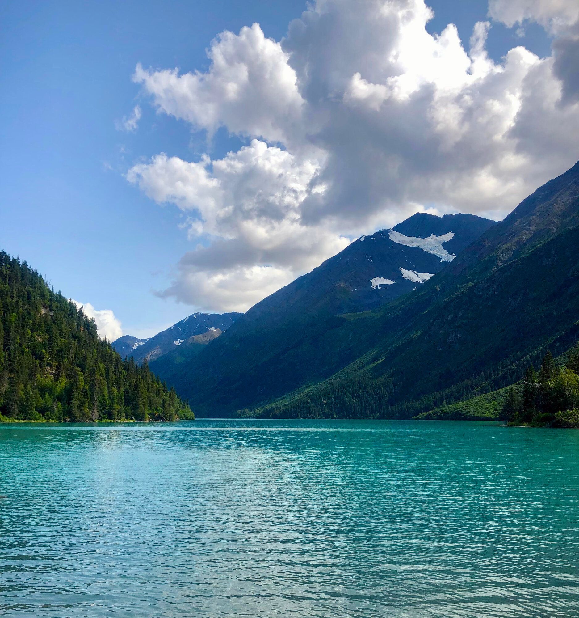 A vibrant turquoise lake nestled between dark green, pine-covered mountains under a blue sky with fluffy white clouds.