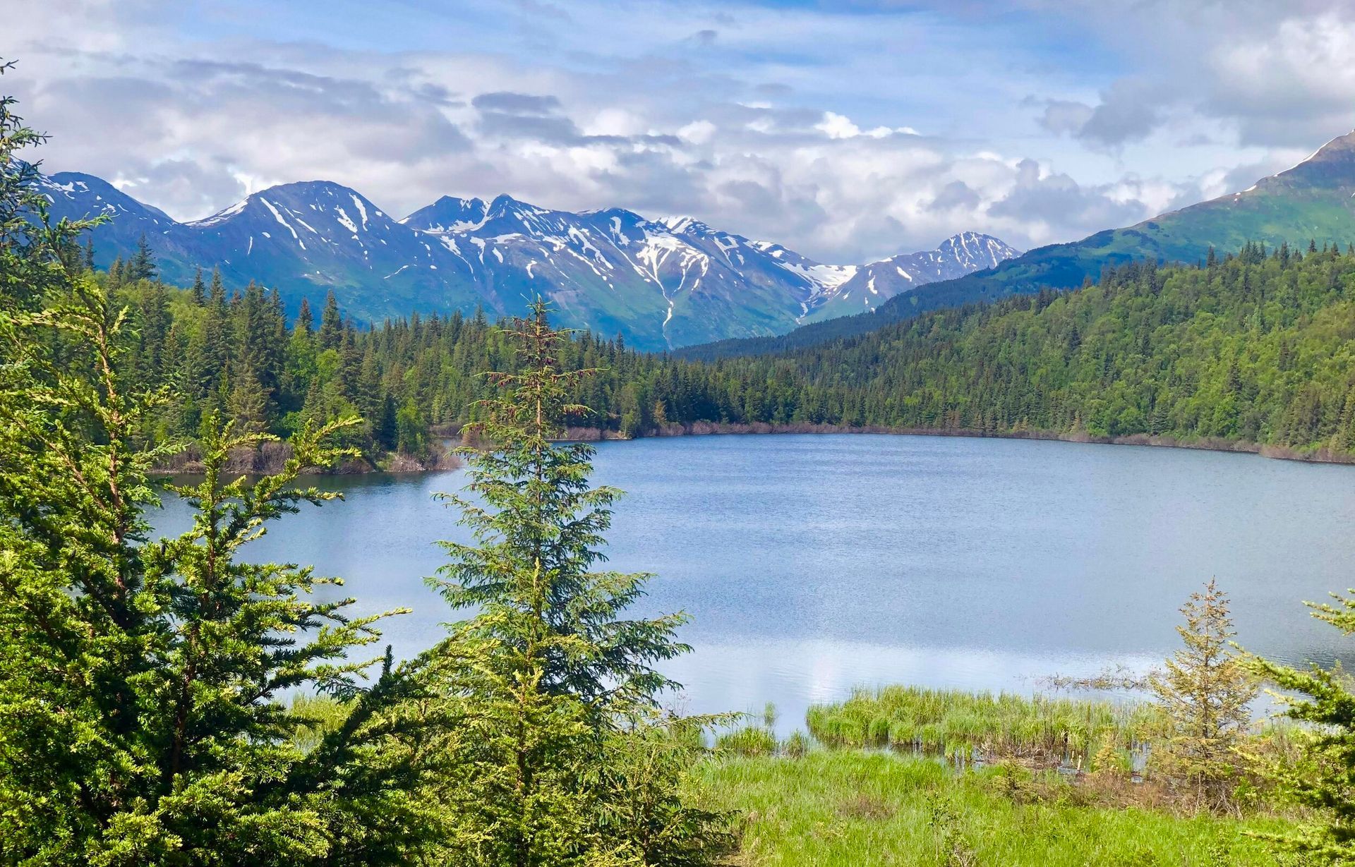 A calm lake nestled in a valley surrounded by dense green pine forests with snow-capped mountain peaks in the distance.