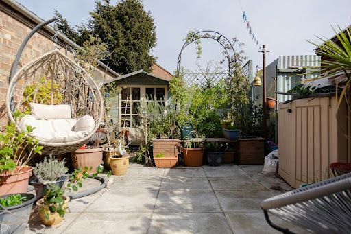 A sunny patio featuring a hanging egg chair, various potted plants, a small garden shed, and a metal archway.