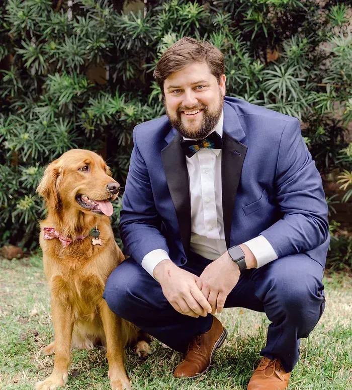 Man in a navy suit crouches beside a golden dog in front of a leafy green wall