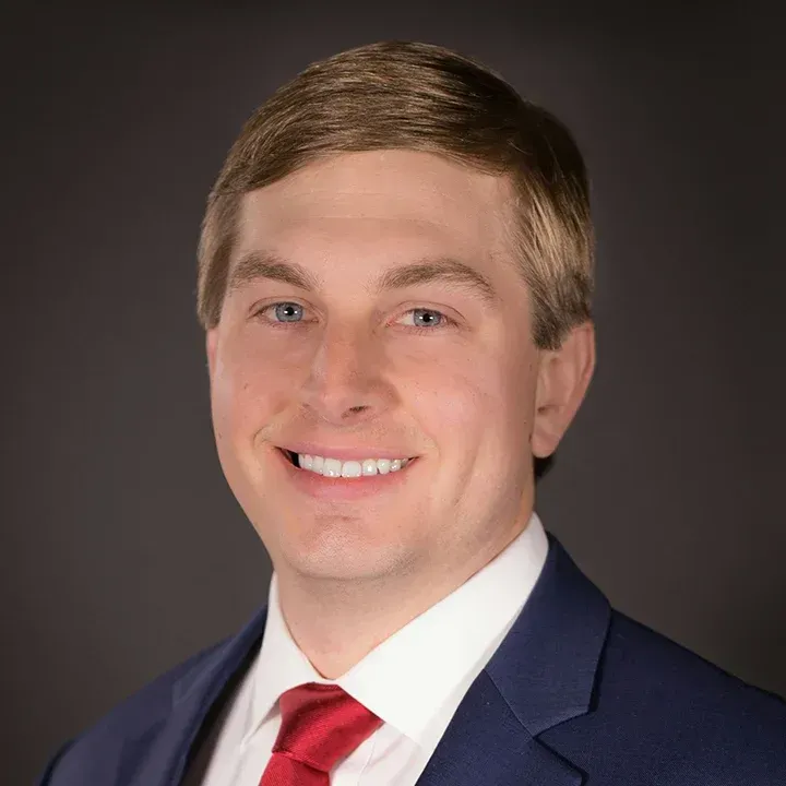 Headshot of a smiling man in a navy suit, white shirt, and red tie on a dark gray background