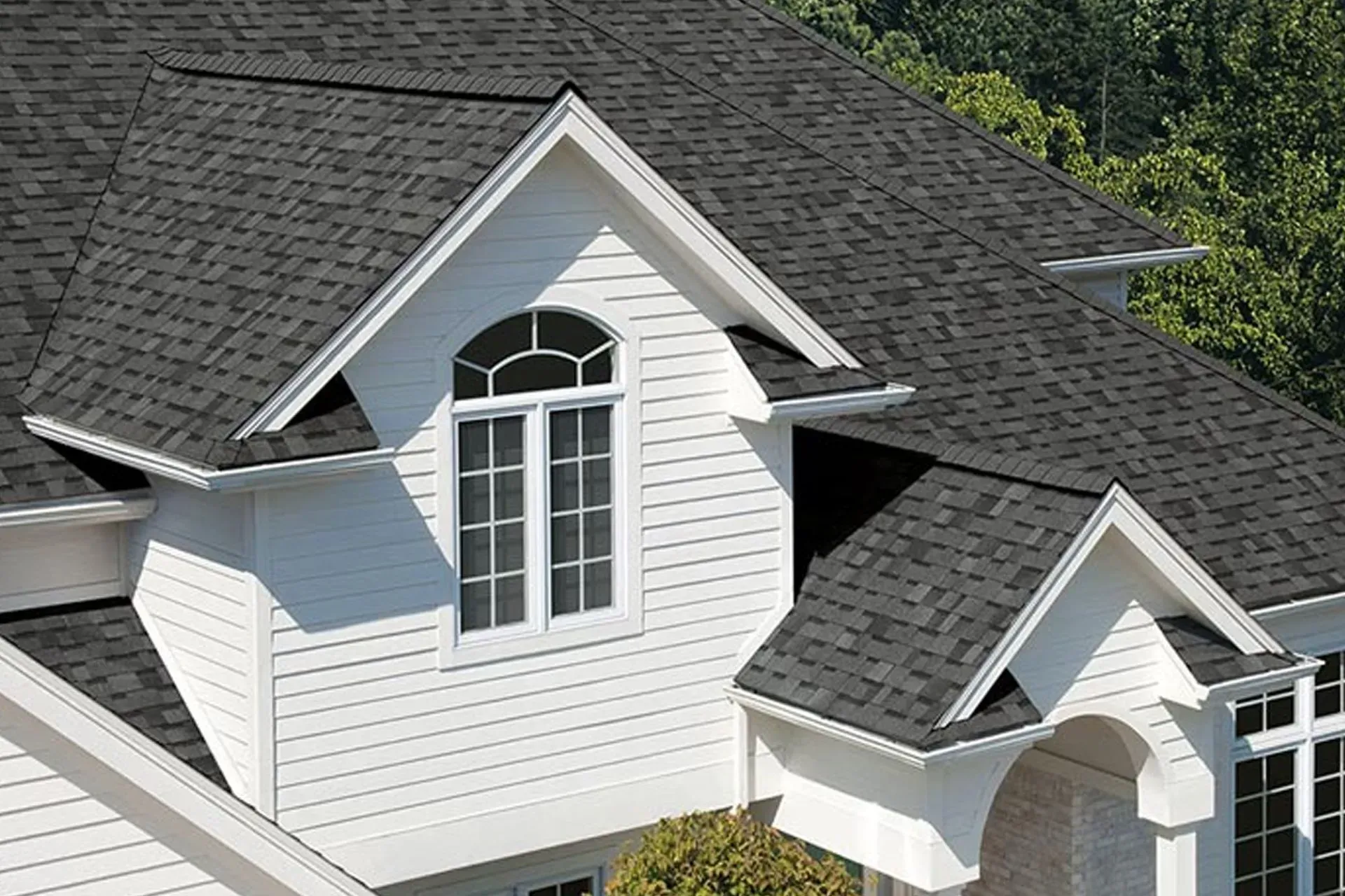White house with dark gray shingle roof and dormer windows against green trees.
