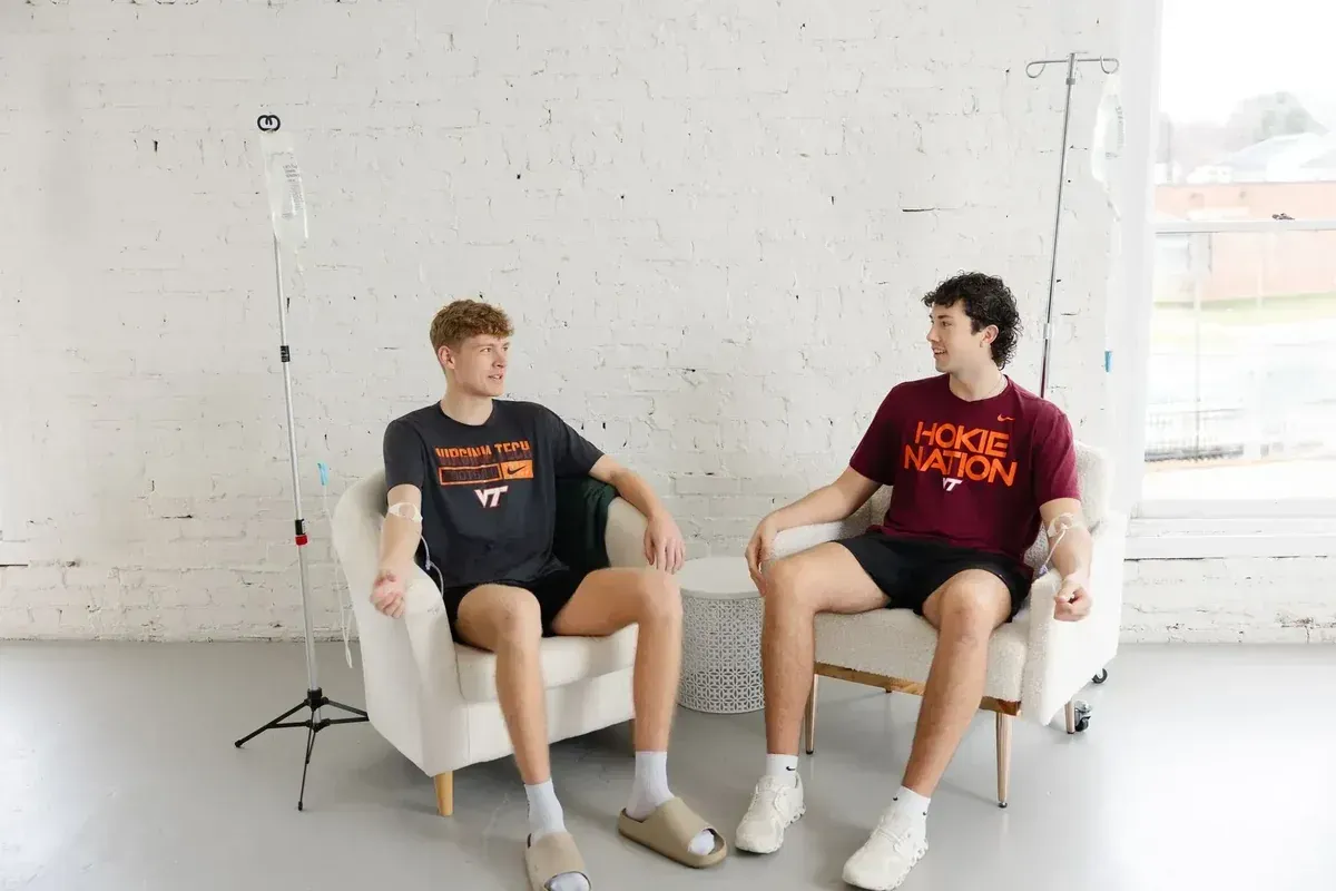 Two people sitting in armchairs in a bright studio with a plain white wall and microphone stand nearby