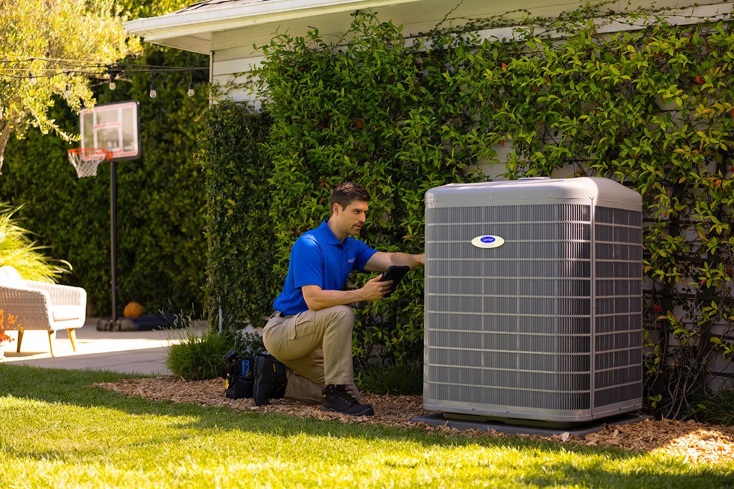 Technician kneeling beside a large outdoor air conditioner unit in a backyard.