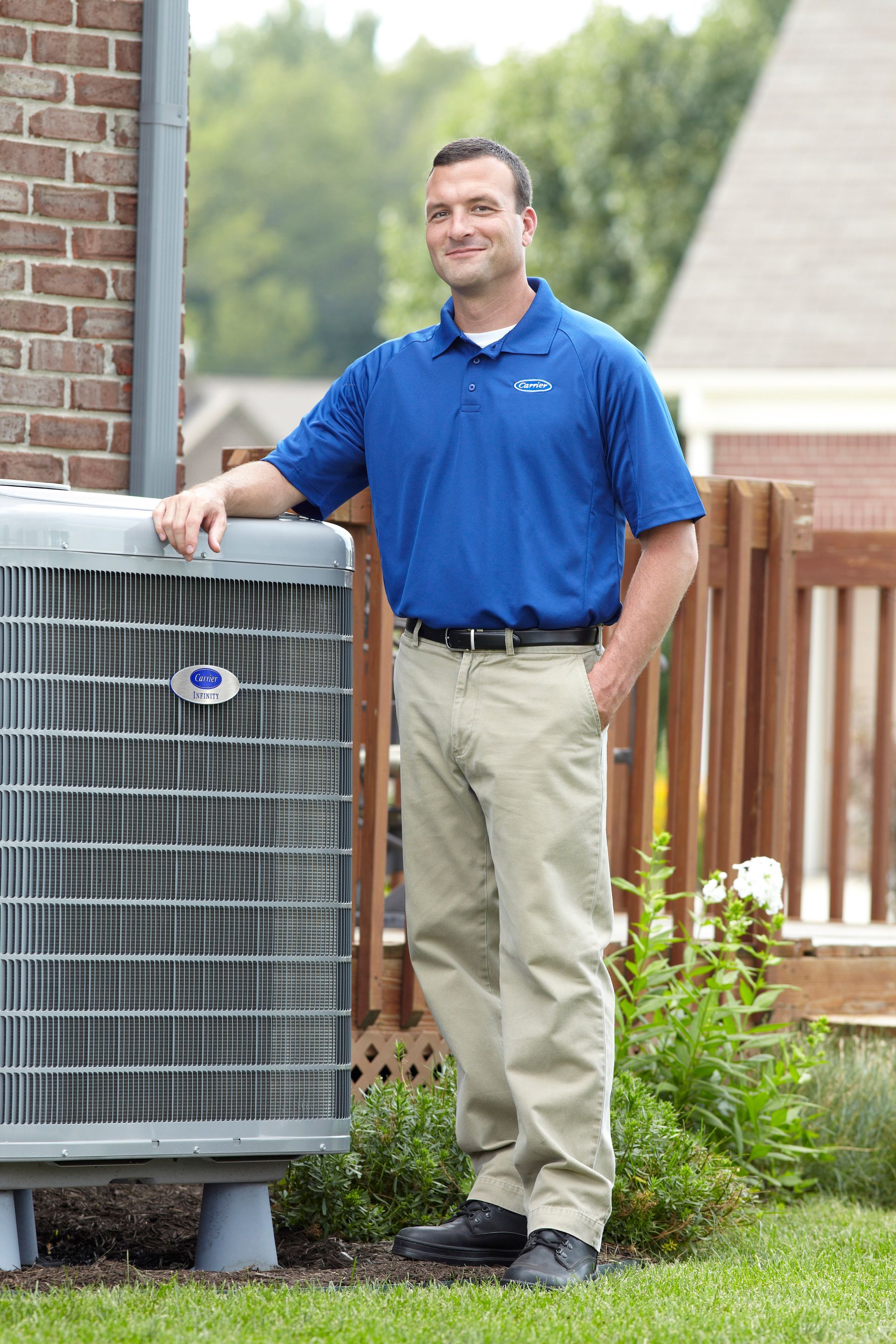 Man standing beside an outdoor air conditioner unit in a backyard, wearing a blue polo and khaki pants.