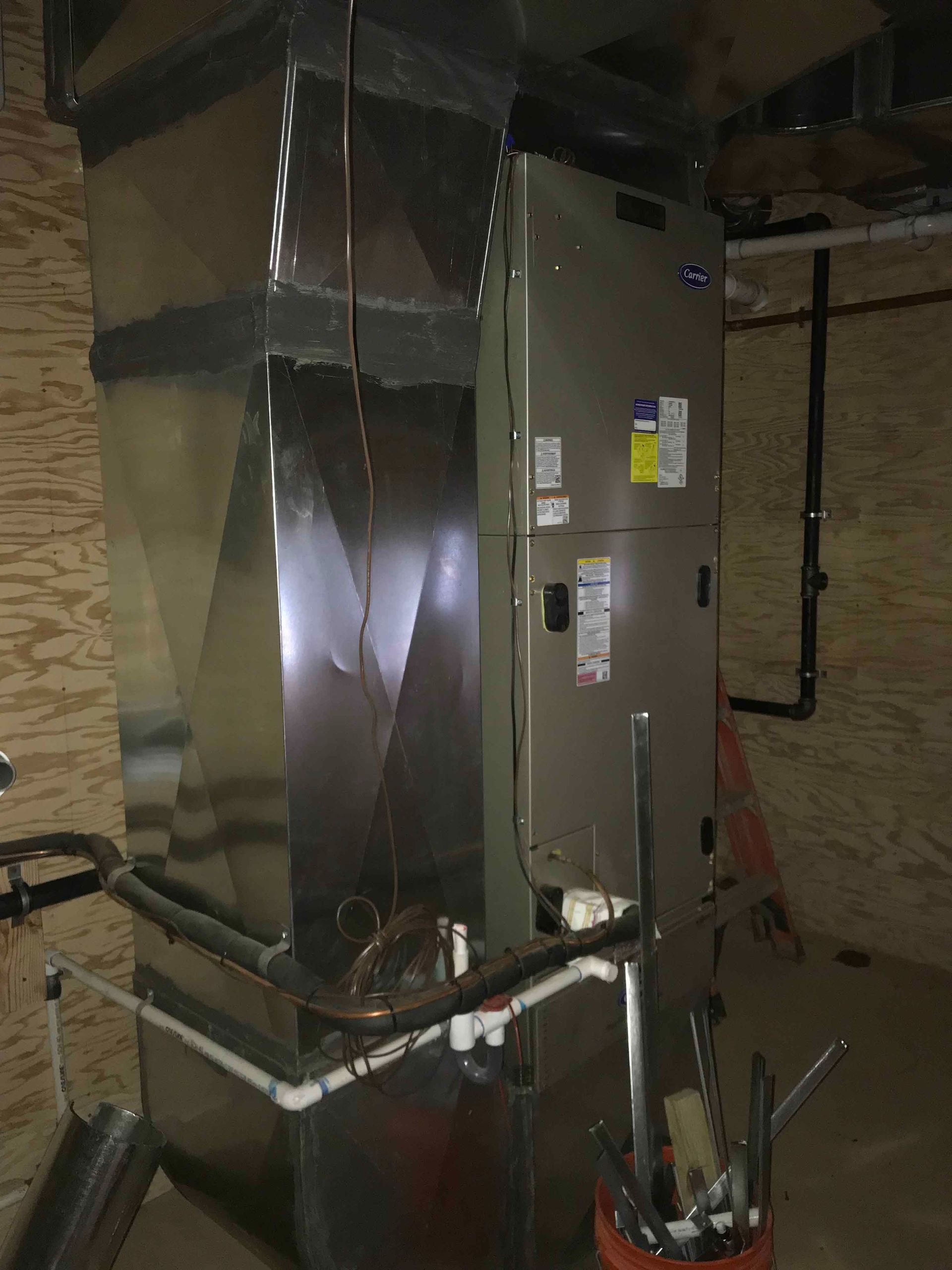 Large HVAC unit in a basement utility room, with metal ductwork and exposed pipes.