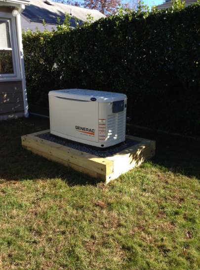 White generator on a raised wooden platform beside a house, in a grassy yard.