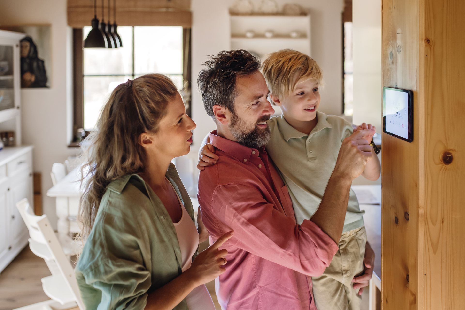 Family using a wall-mounted tablet in a bright kitchen, with one adult and two children looking at the screen