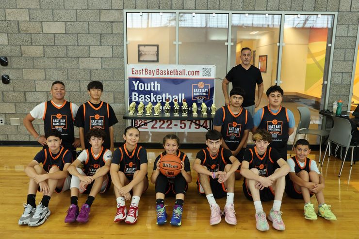 A basketball team poses for a group photo on a wooden indoor court at the East Bay Basketball training center.