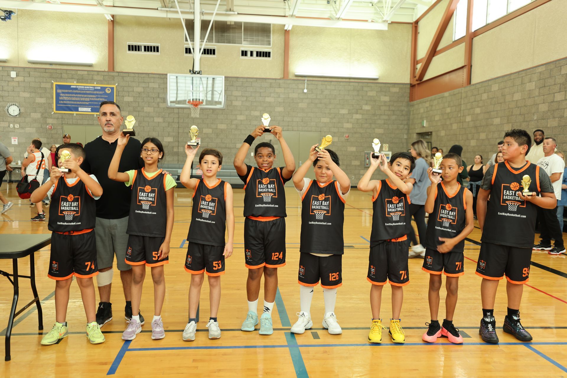 A basketball coach and eight players in team jerseys holding trophies on an indoor court.