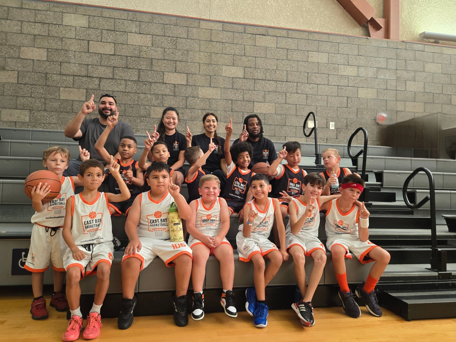 A basketball team in matching orange and white uniforms poses on gymnasium bleachers, with coaches standing behind.