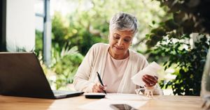 Person writing at a desk with a laptop and papers in a bright indoor garden setting Person writing at a desk with a laptop and papers in a bright indoor garden setting