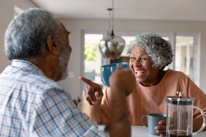 Two people smiling and talking at a kitchen table with mugs and a French press Two people smiling and talking at a kitchen table with mugs and a French press